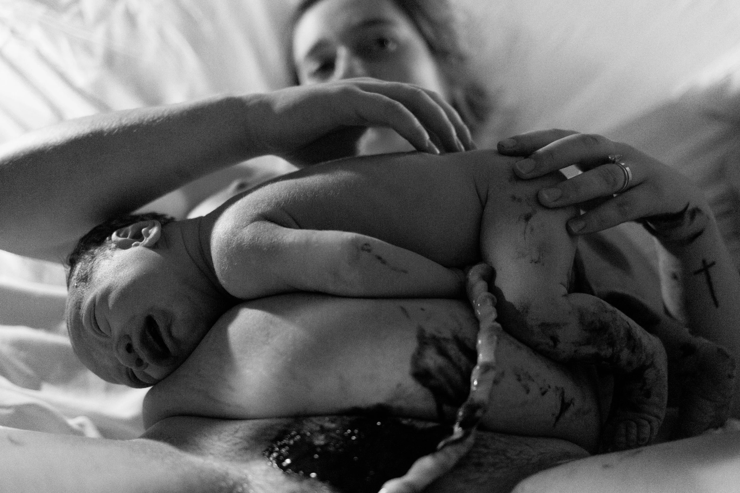 A black and white photo of a woman holding a newborn baby close to her face, both in bed. The baby appears to be resting or sleeping, and the woman has tattoos on her hands and a ring on her finger.