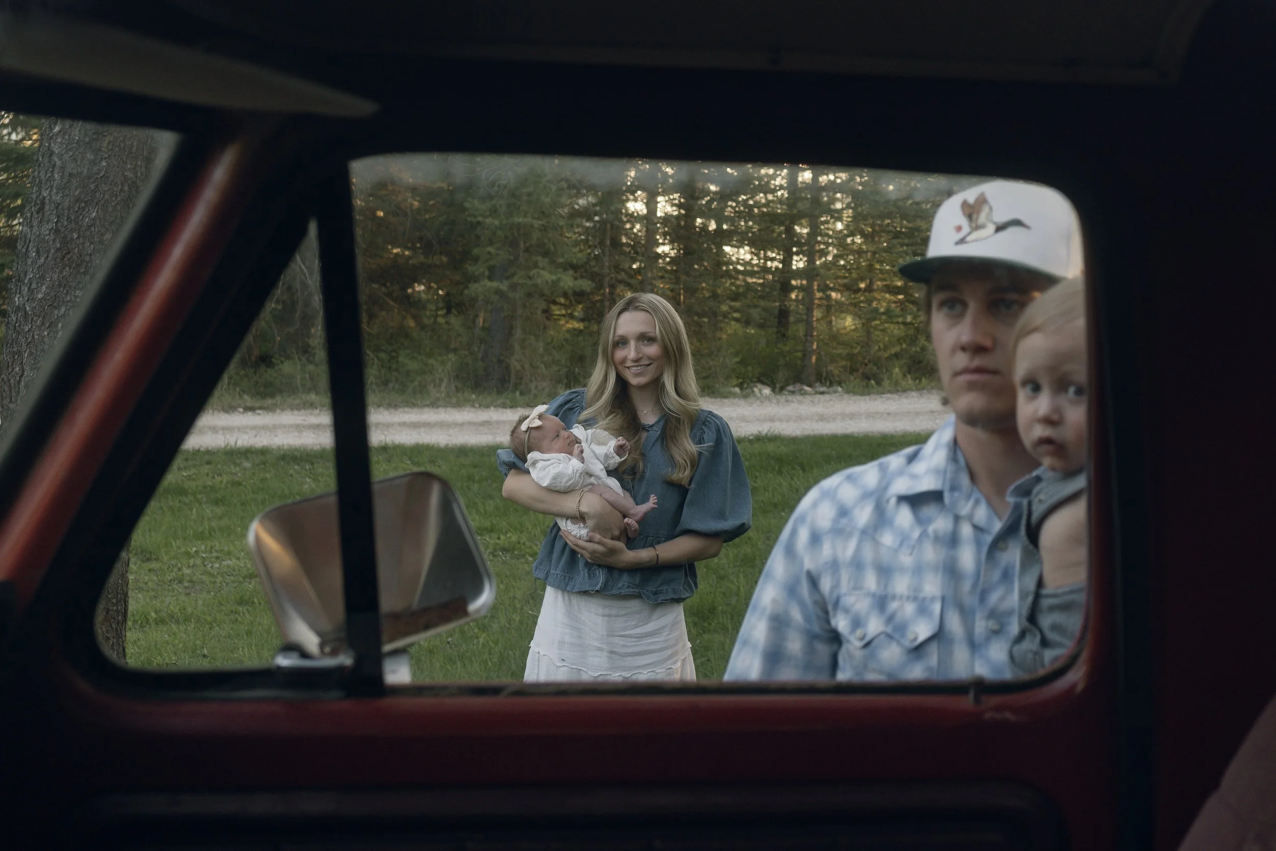 A woman with long blonde hair holding a baby standing outside in front of a forest, while two men look on from inside a vehicle. The image appears to be viewed through the vehicle's side mirror.