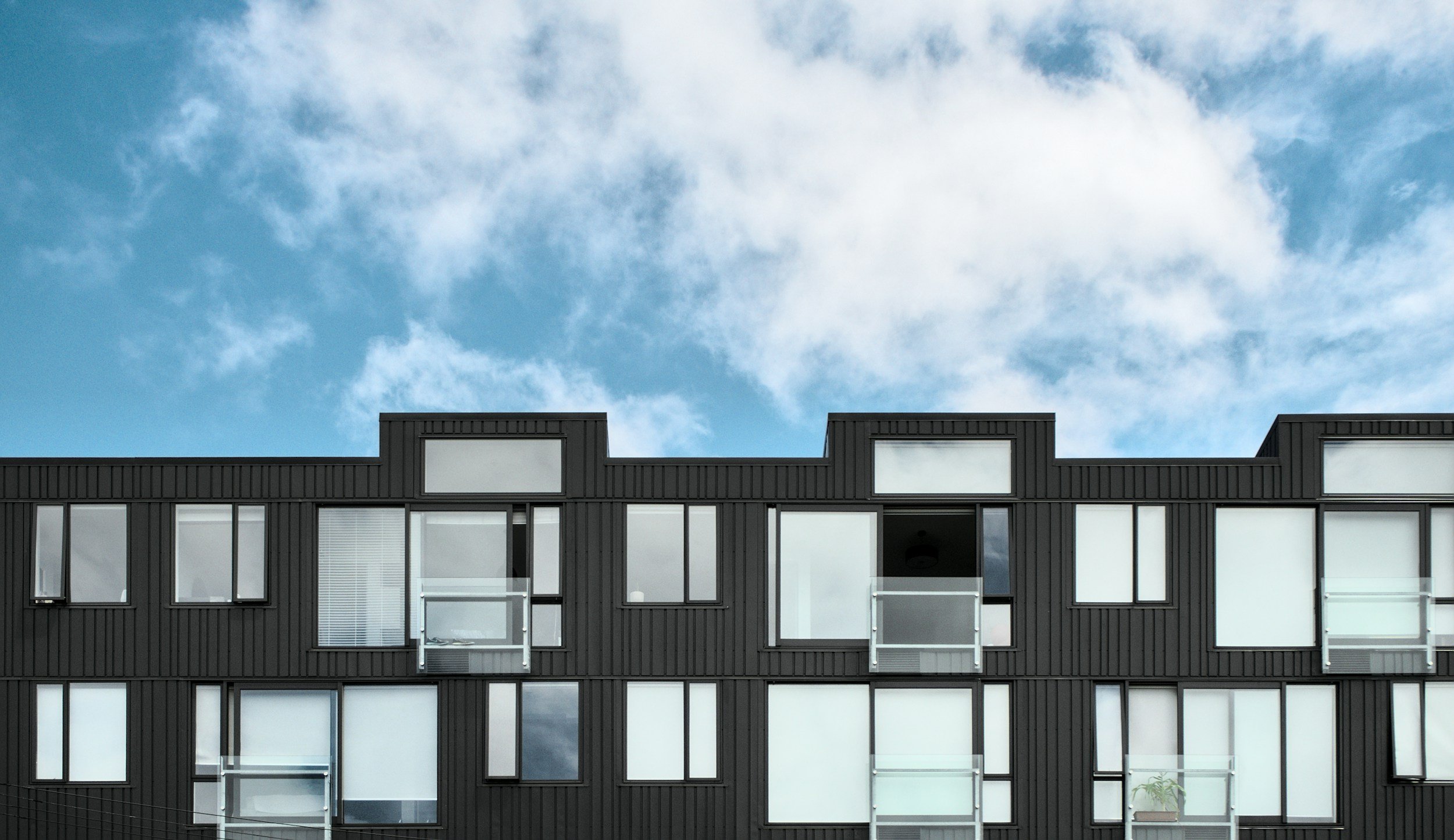 Modern black apartment building with multiple windows and small balconies, against a cloudy blue sky.