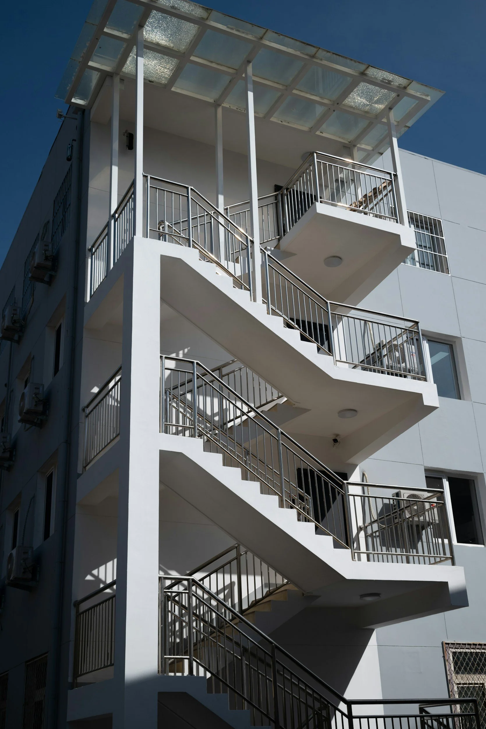Exterior of a modern white multi-story building with a metal staircase and glass rooftop cover, against a clear blue sky.