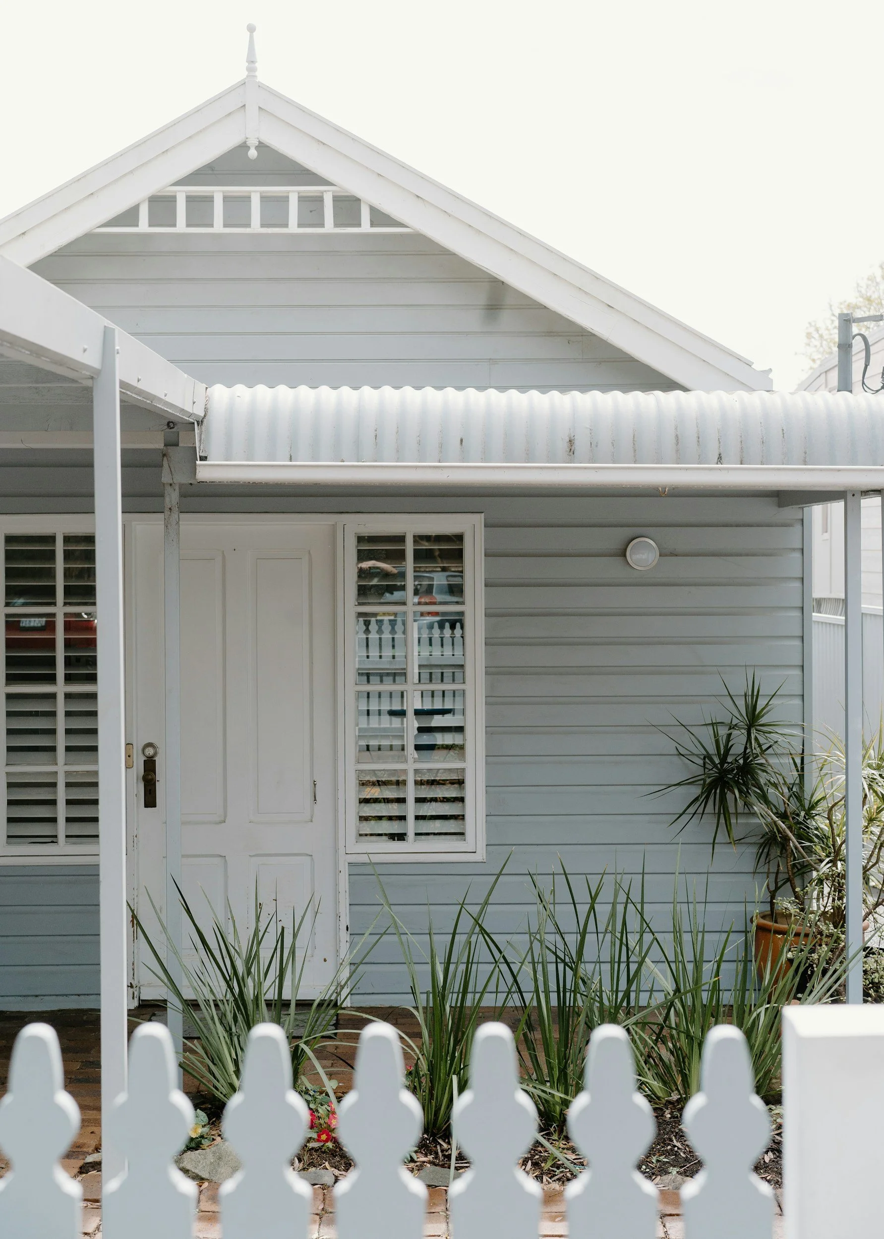 Front view of a light blue house with a white door, a window with white shutters, and a white picket fence in the foreground. There are plants in pots and some greenery outside.