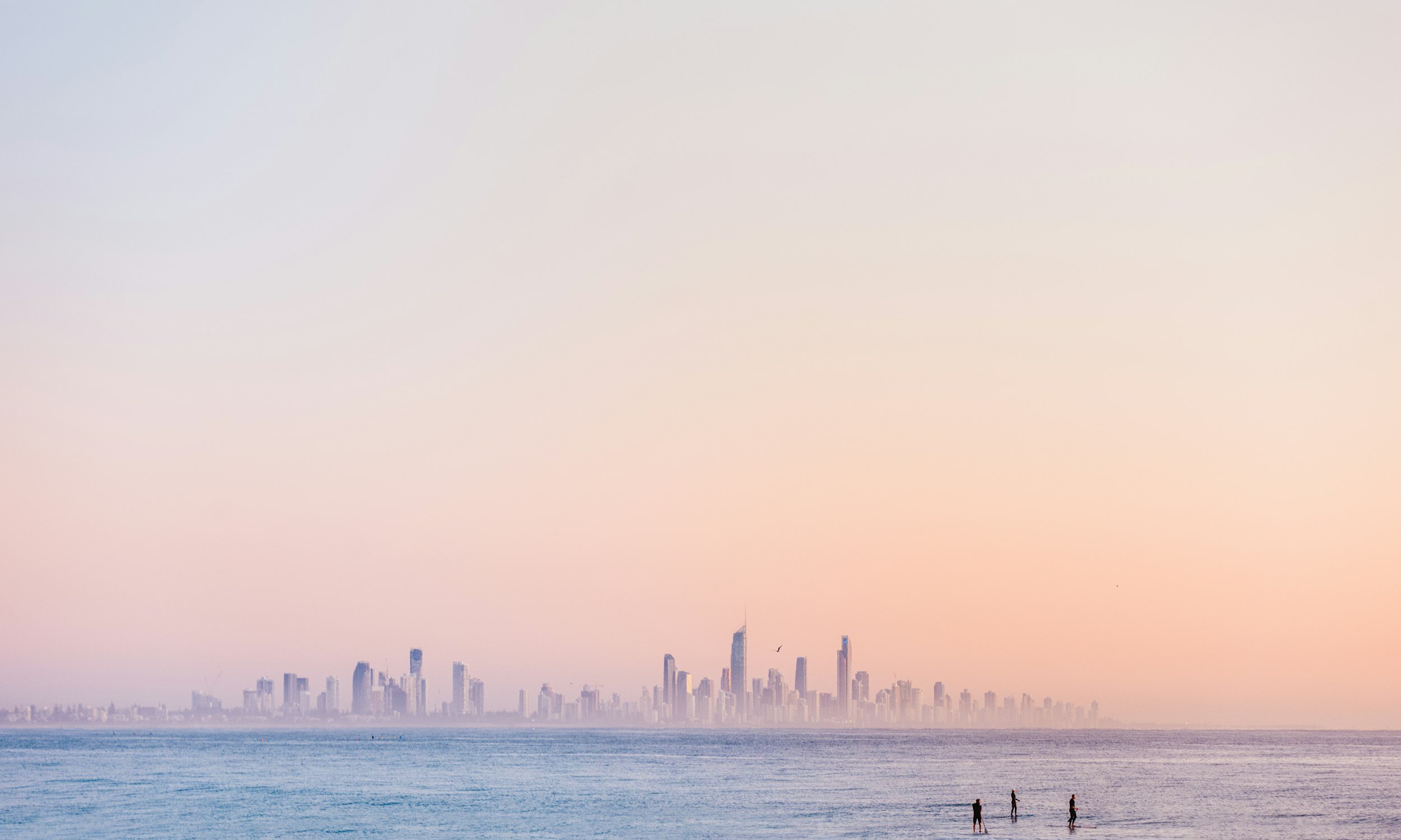 A city skyline on the horizon seen across calm water during sunset, with three people standing in shallow water in the foreground.