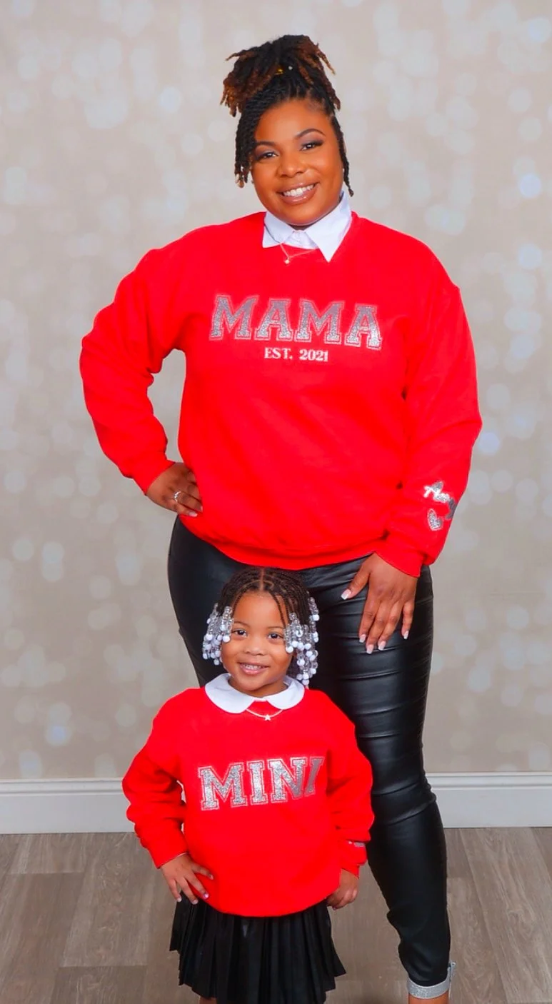 An adult woman and a young girl standing together, both wearing matching red sweaters with silver lettering. The woman's sweater says "MAMA EST. 2021" and the girl's sweater says "MINI." The woman has her hair styled in twists and is smiling, while t