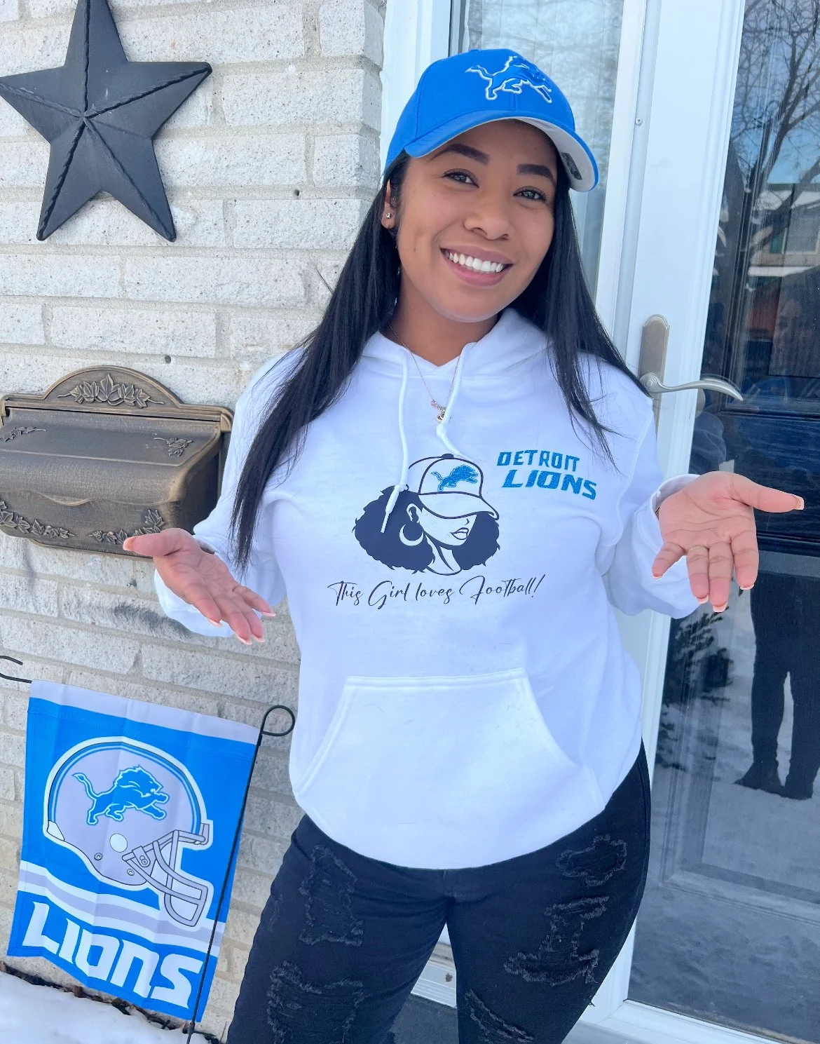 A woman wearing a Detroit Lions hoodie and cap, standing outside near a glass door, smiling with her hands raised in a shrug gesture.