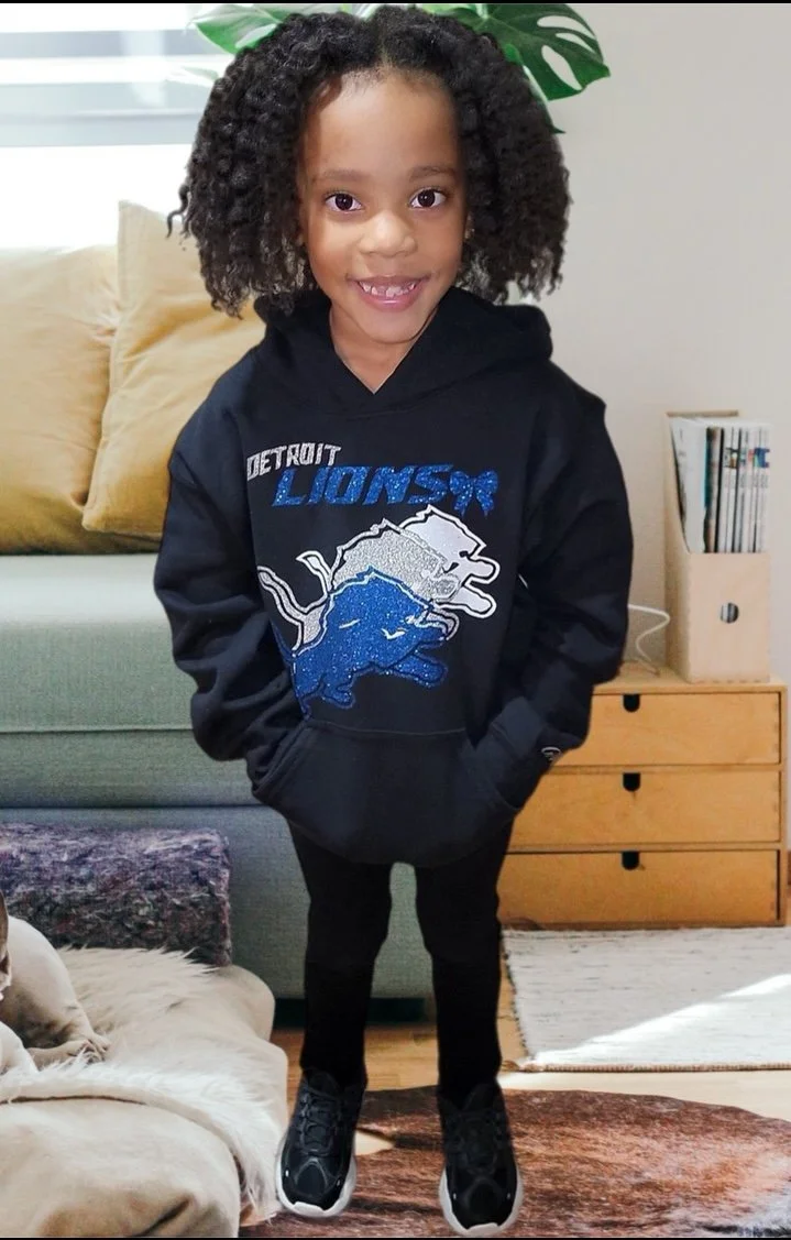 A young girl with curly hair smiling, wearing a black Detroit Lions hoodie, standing indoors with a sofa, books, and a plant in the background.