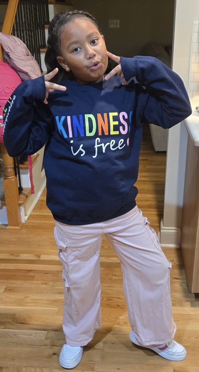 A young girl in a navy blue sweatshirt with rainbow-colored letters spelling 'KINDNESS is free,' posing indoors with her hands near her face making a peace sign. She has dark hair pulled back and is wearing light-colored cargo pants and white sneaker