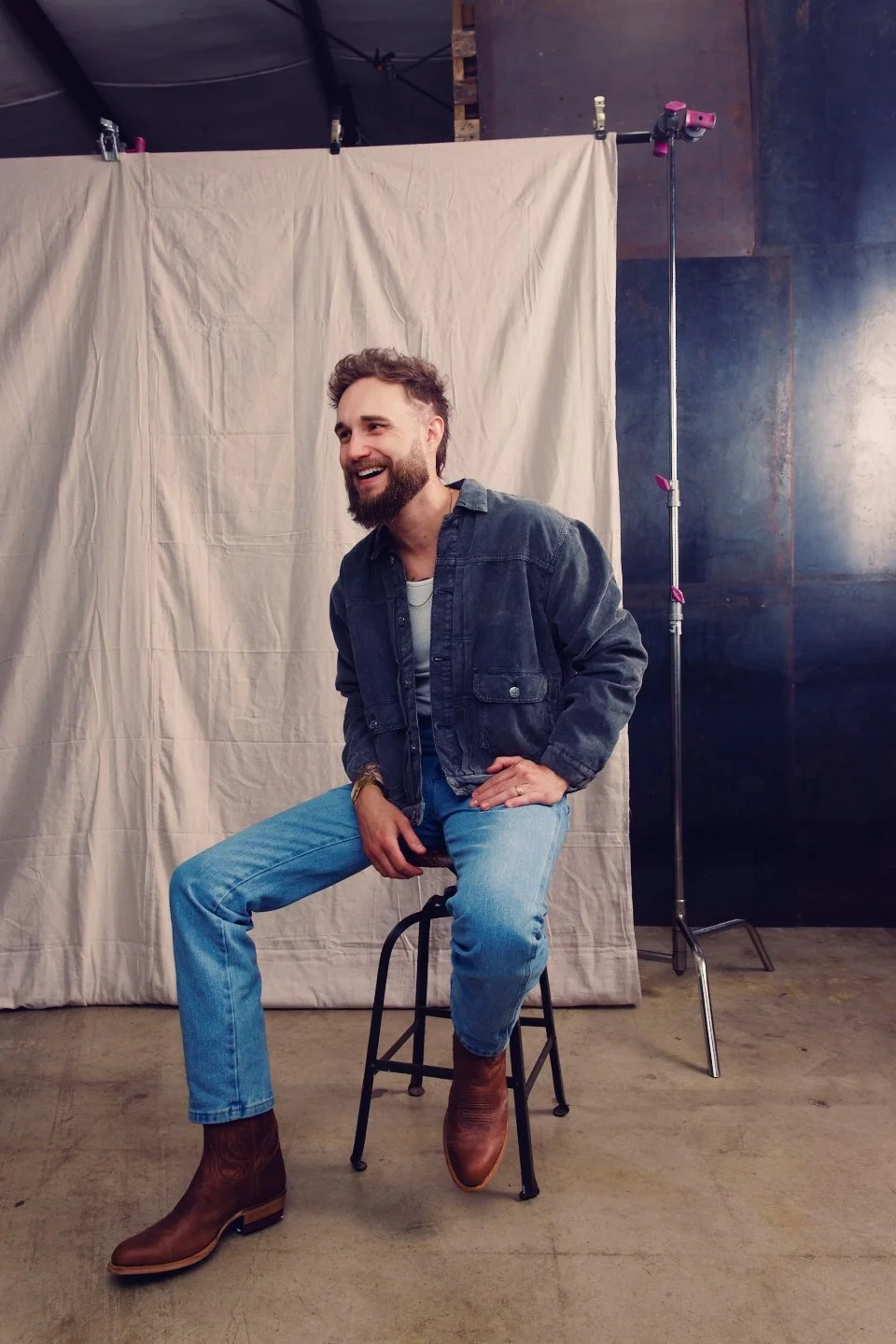 A man with a beard and curly hair smiling and sitting on a stool in front of a beige cloth backdrop, wearing a denim jacket, white shirt, blue jeans, and brown cowboy boots, with photo studio equipment visible.