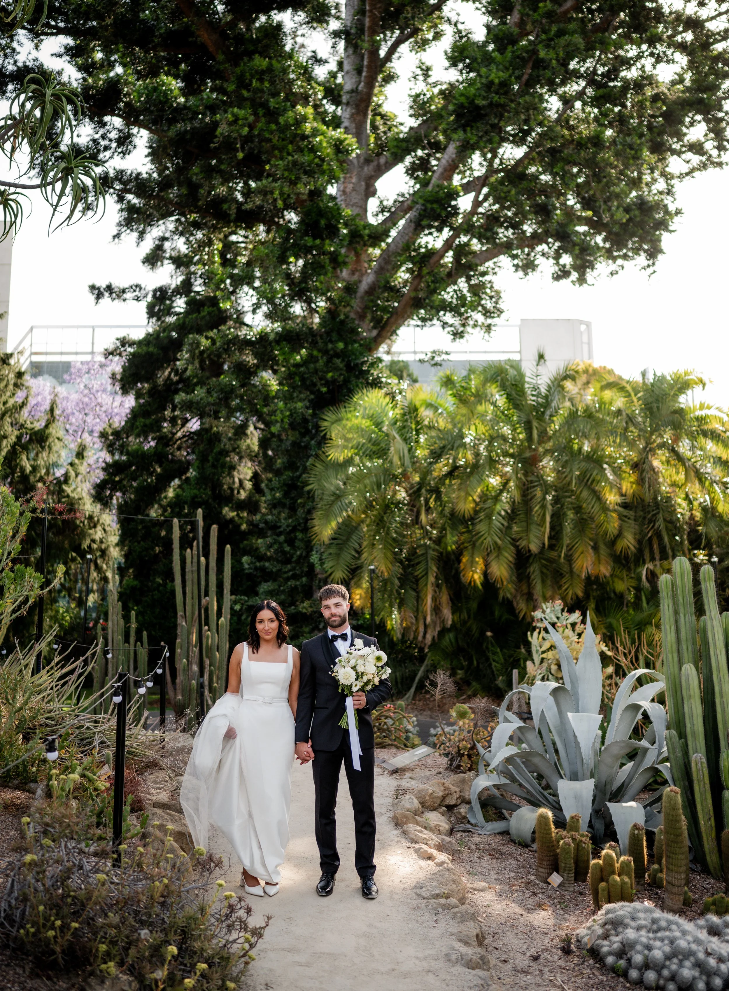 A bride and groom walk hand in hand through a lush, green Adelaide botanical gardens on their wedding day.