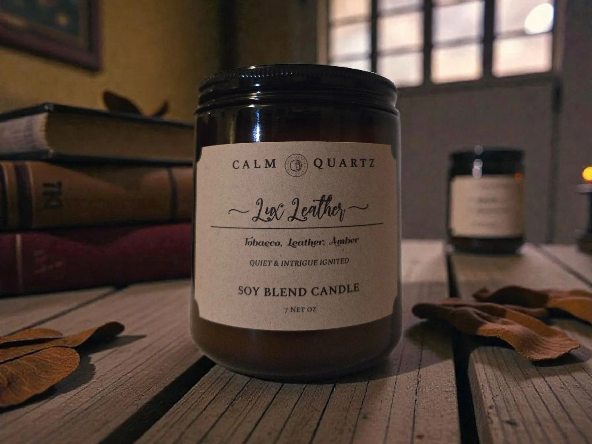 lux leather candle surrounded by dried tabacco leaves on a wooden table next to a stack of leather bound books