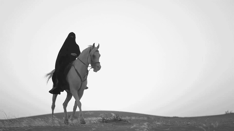 A person wearing a black cloak riding a horse in a desert landscape with a clear sky.