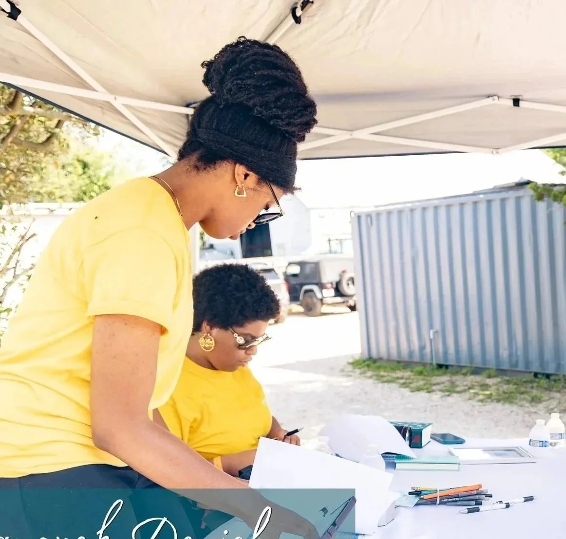 Two women wearing yellow shirts working outdoors under a tent with supplies and papers on a table.