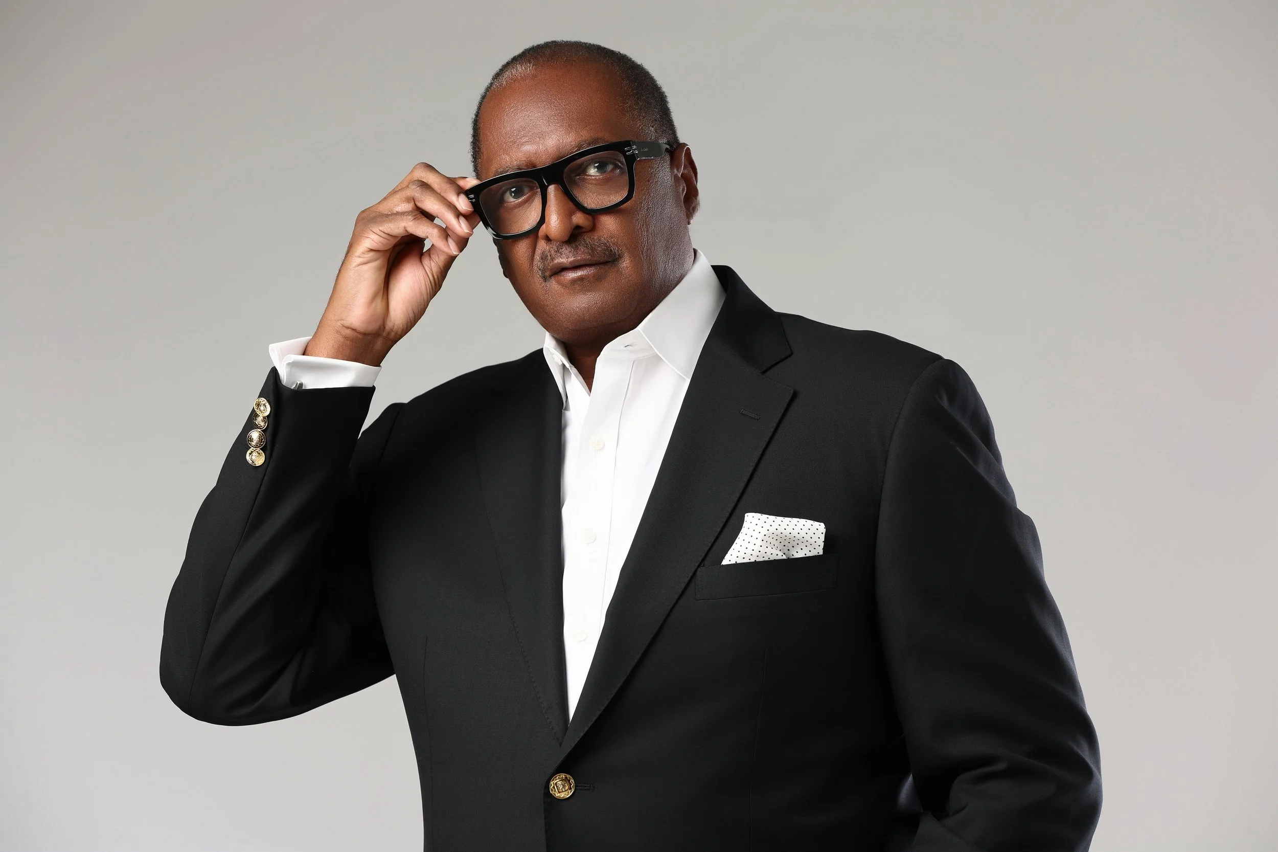 Close-up portrait of a middle-aged man with short hair, wearing black glasses, a white dress shirt, a dark gray suit, and a white pocket square, against a plain white background.