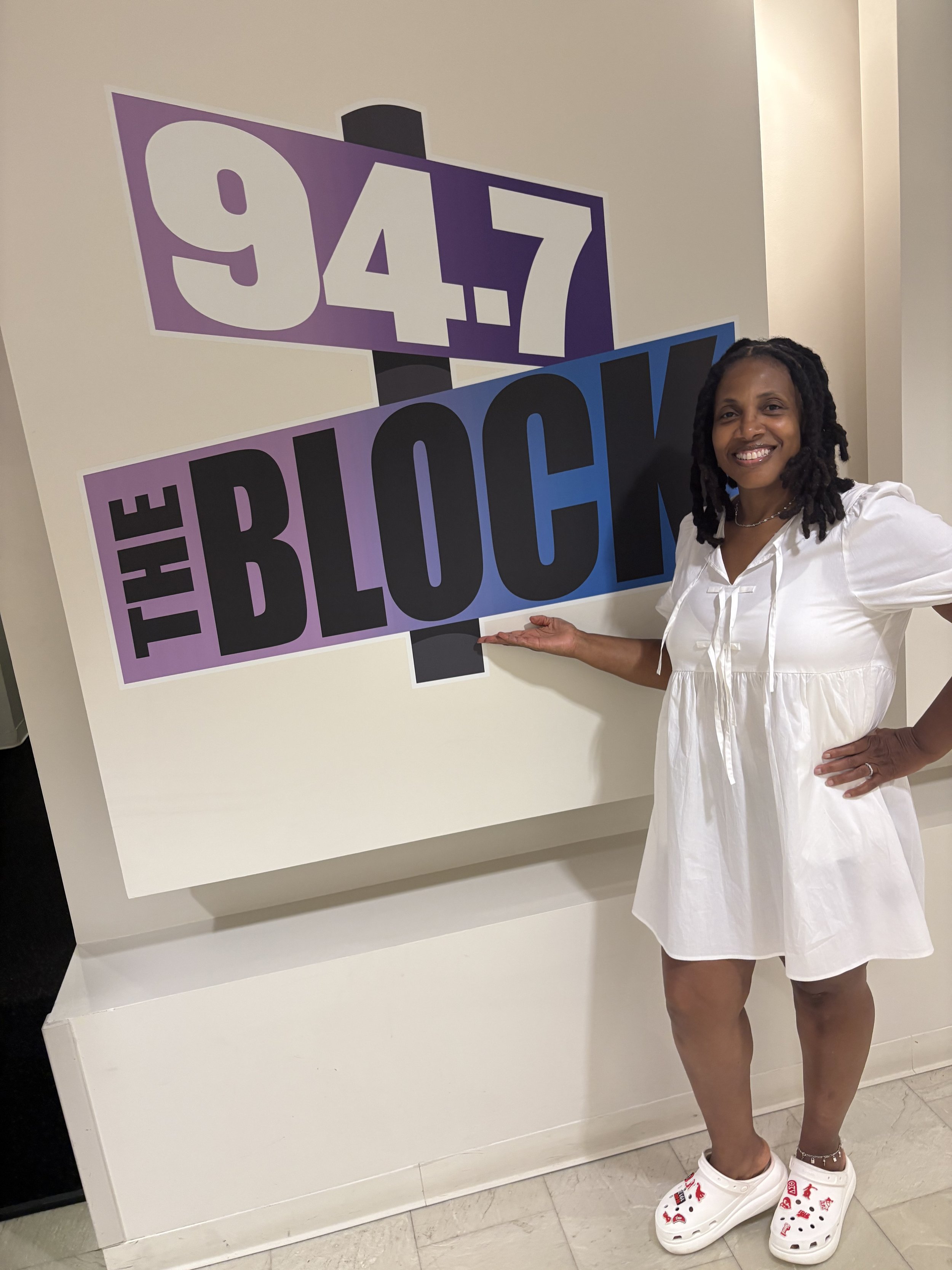 A woman in a white dress smiling and pointing at a sign that reads '94.7 THE BLOCK' in purple, black, and blue letters.