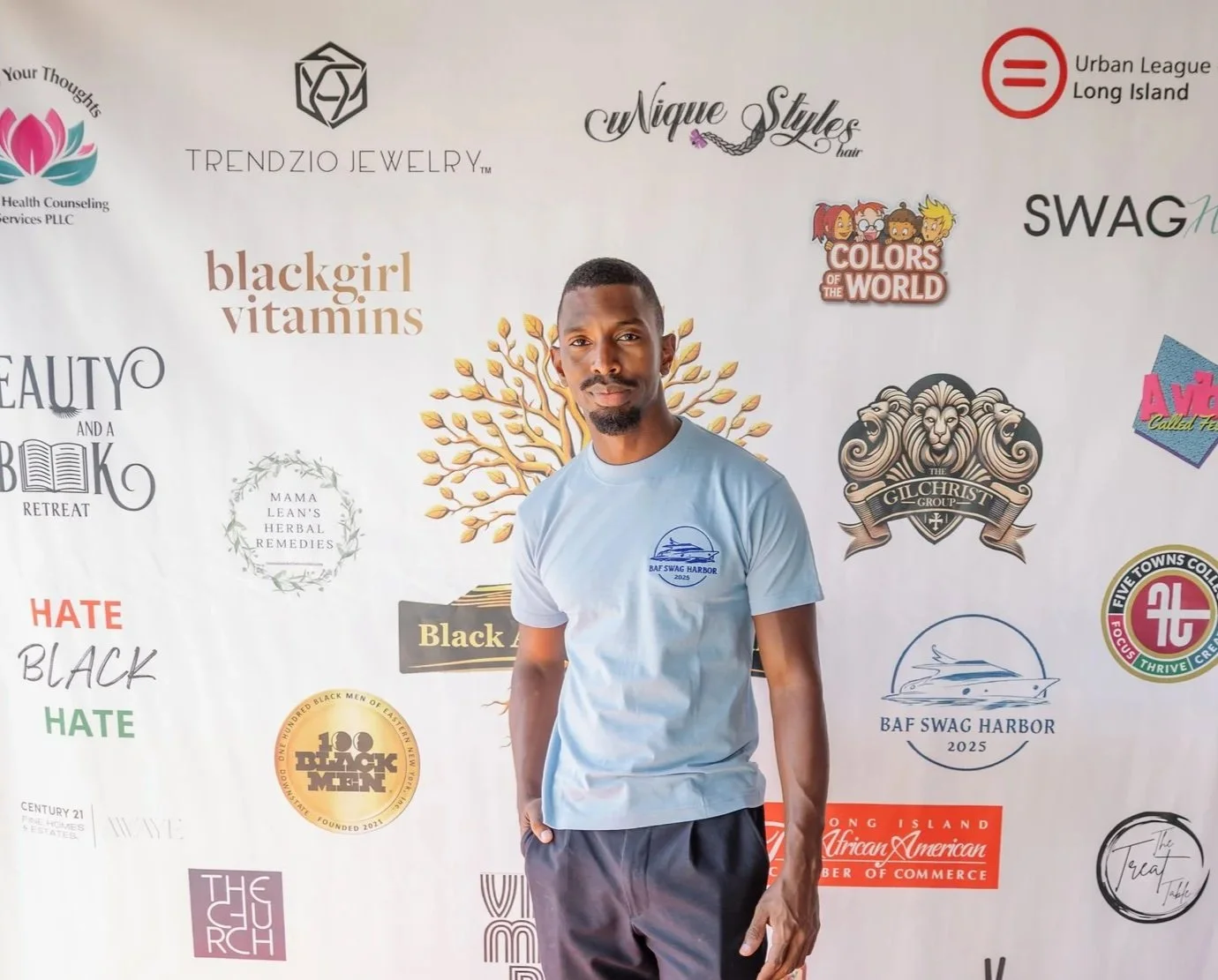 Man standing in front of a backdrop with various logos and texts, wearing a light blue t-shirt and dark pants.
