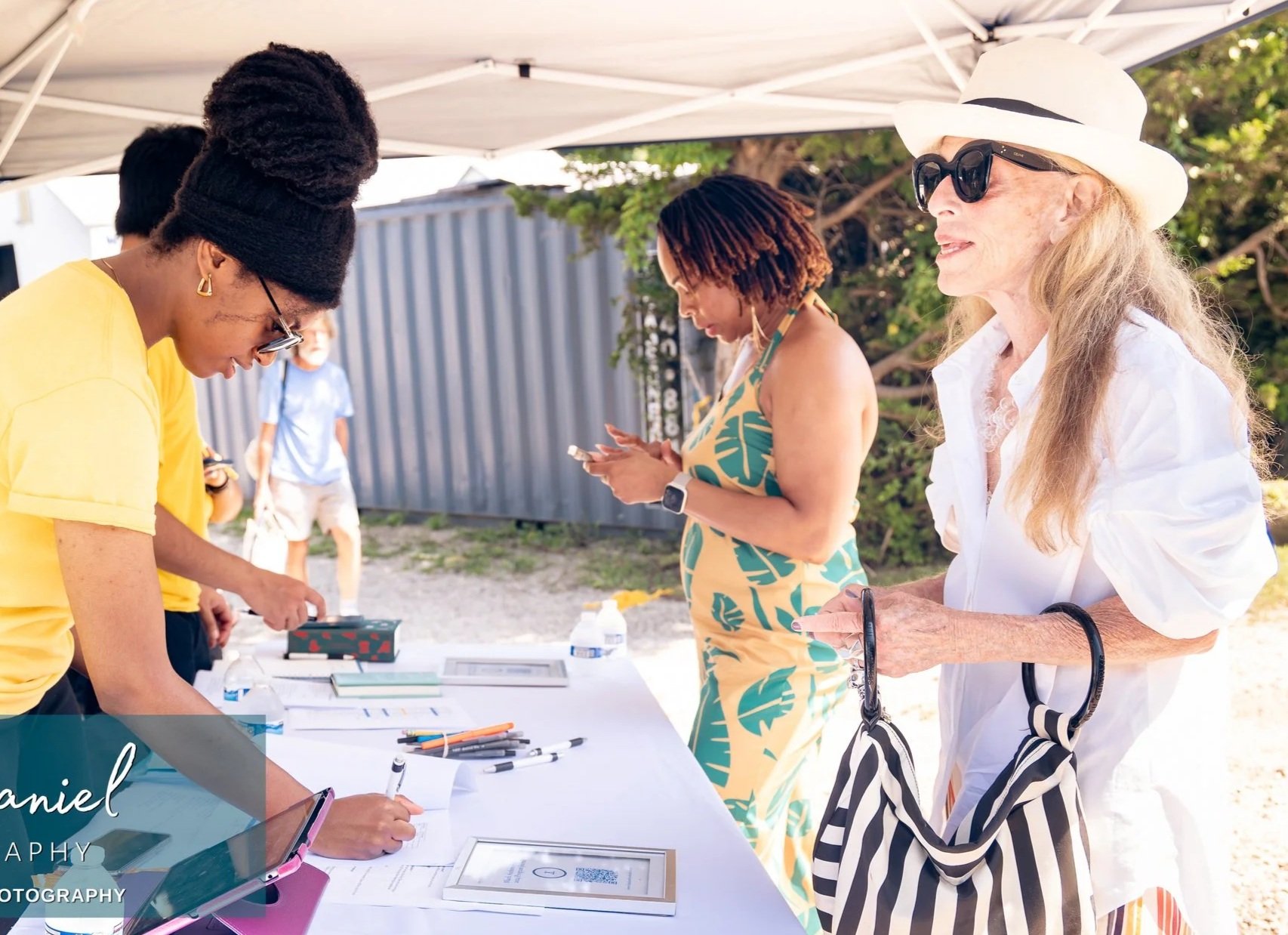 Women and people at an outdoor registration table under a white canopy during a sunny day.