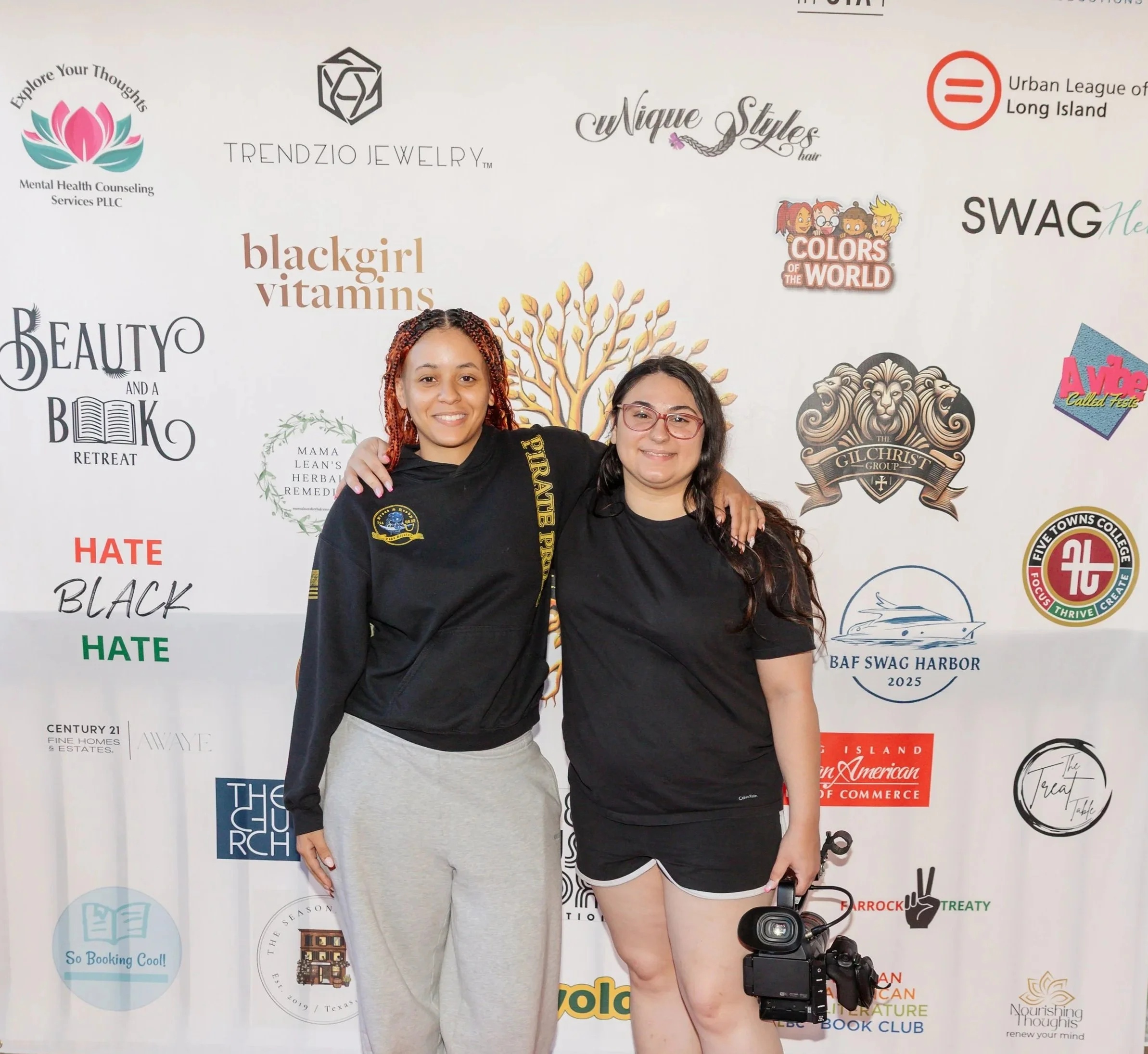 Two young women standing in front of a backdrop with various logos. The woman on the left has red braids and is wearing a black sweatshirt and gray sweatpants. The woman on the right has long dark hair, glasses, and is wearing a black t-shirt and black shorts, holding a camera.