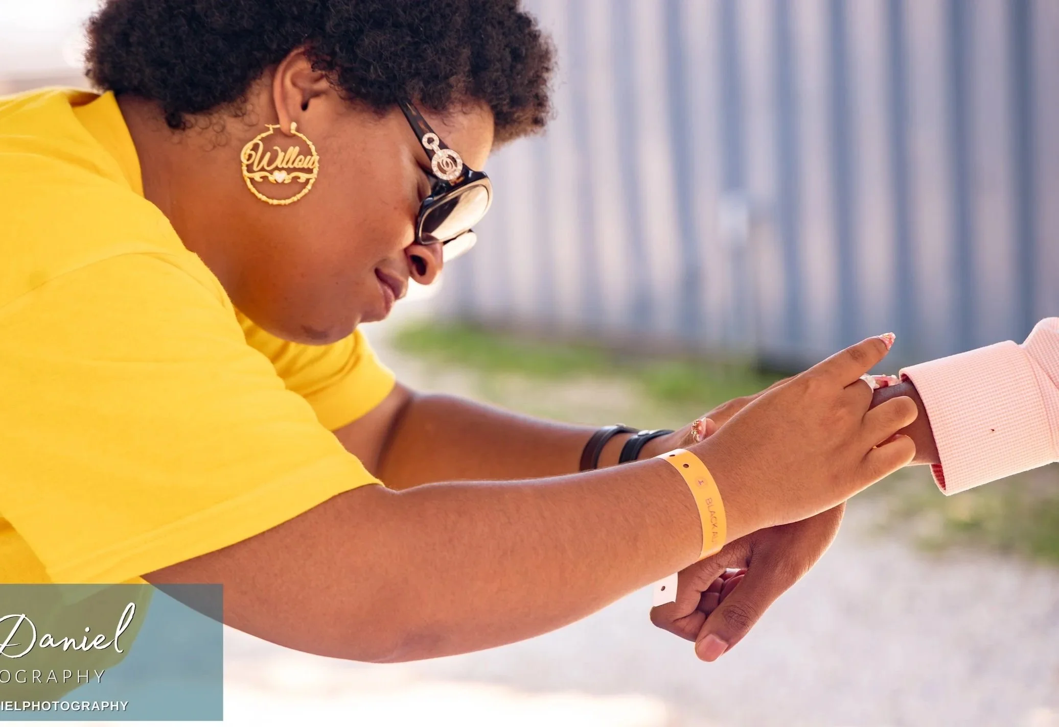 A woman wearing a yellow T-shirt, glasses, and a large 'Black Lives Matter' earring touches her wrist, which has a wristband, while her hand is being gently held by someone wearing a pink sleeve.