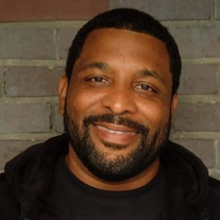 Close-up of a man with a beard smiling, wearing a black hoodie, standing against a brick wall.
