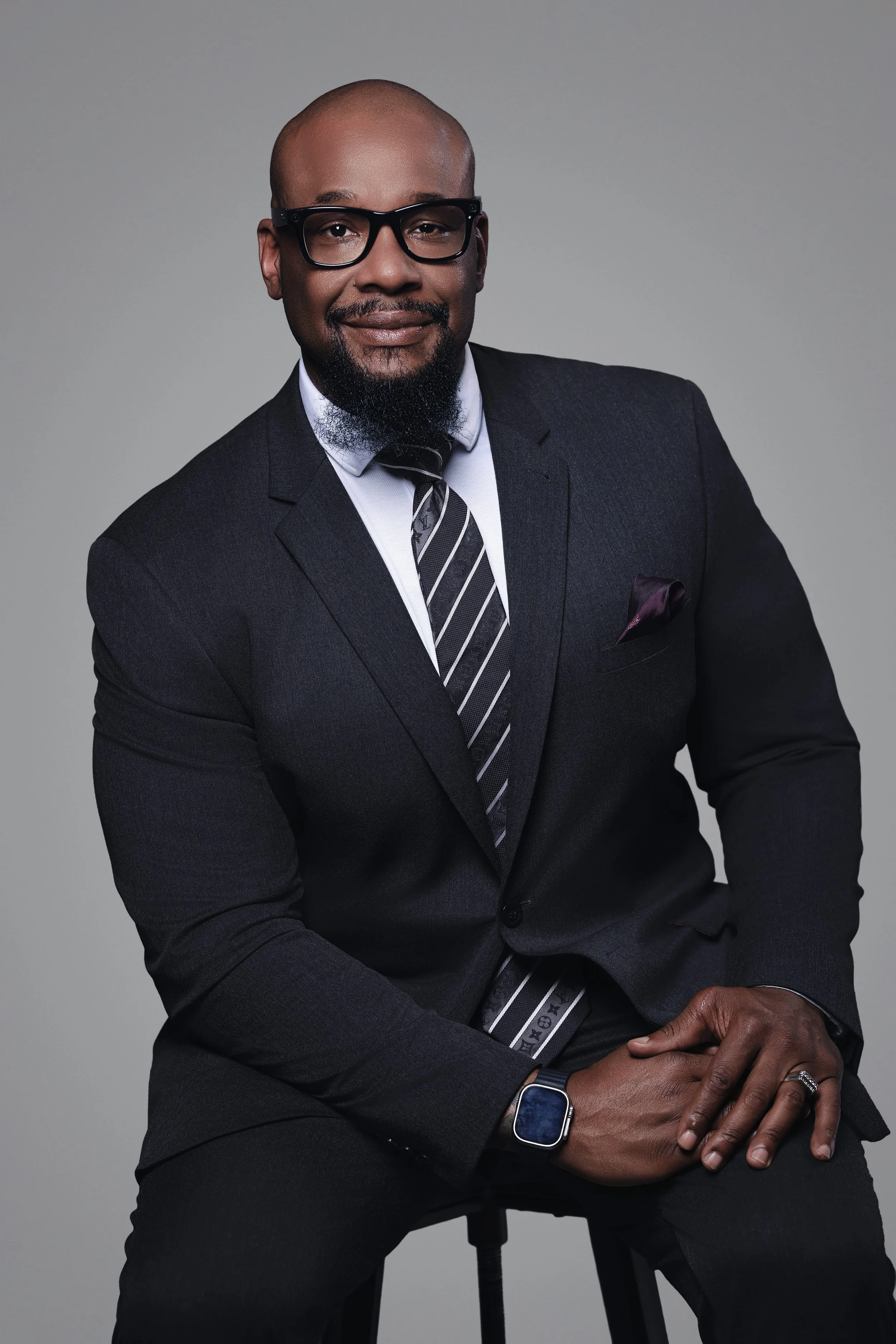 A well-dressed man in a black suit with a striped tie, glasses, and a smartwatch sitting on a black stool against a plain gray background.