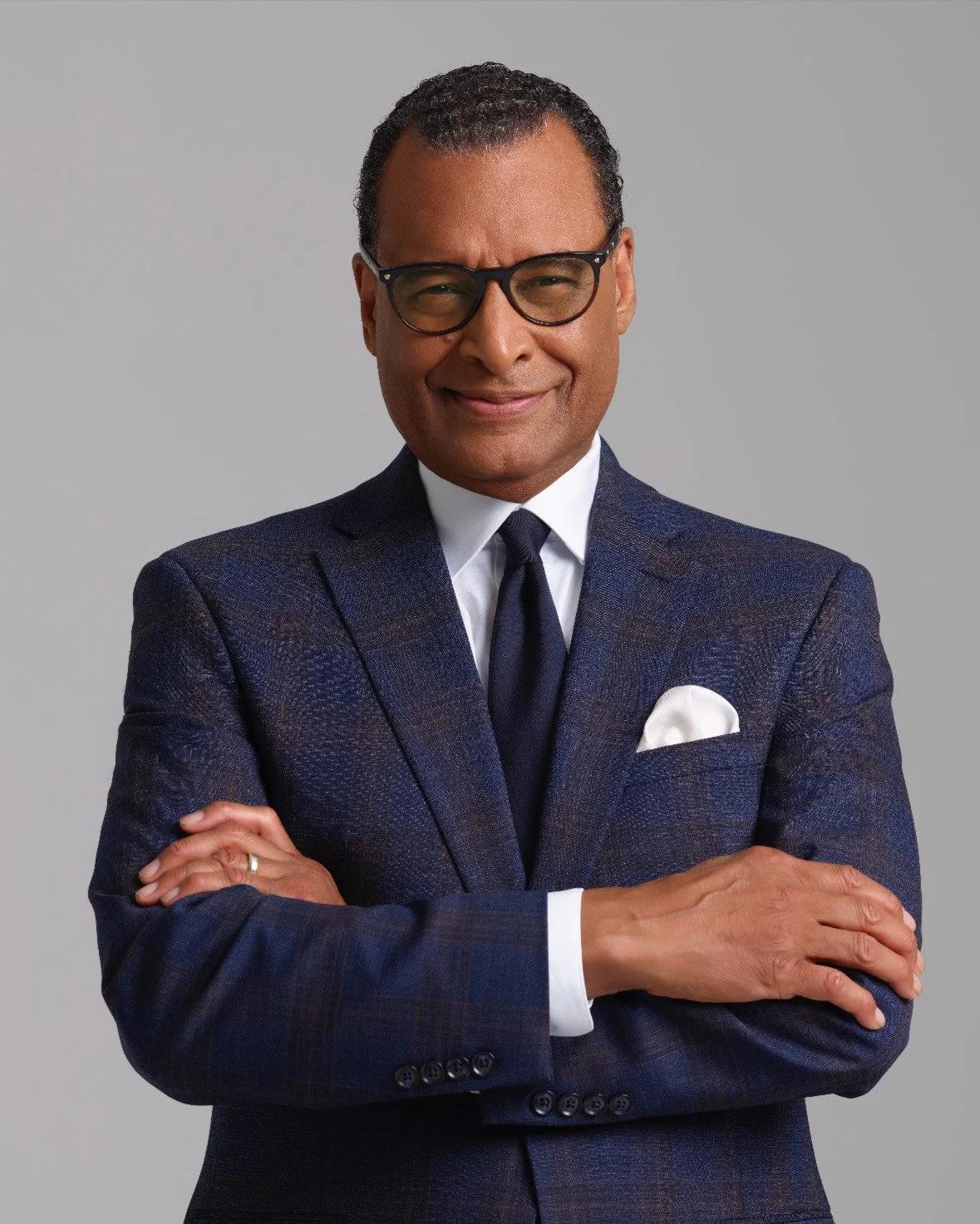 A confident man wearing a navy plaid suit, white shirt, navy tie, and black glasses, standing against a plain gray background with his arms crossed and a slight smile.