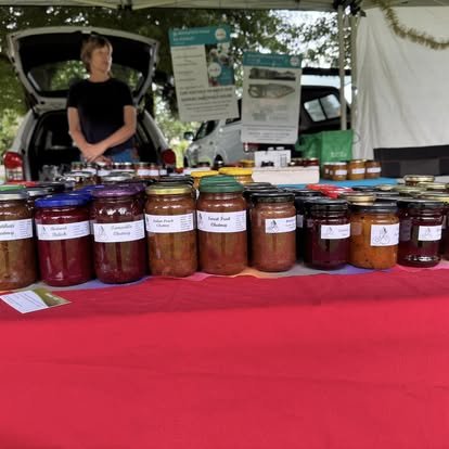 Assorted jars of jams and chutneys displayed on a table with a red tablecloth at an outdoor market stall.