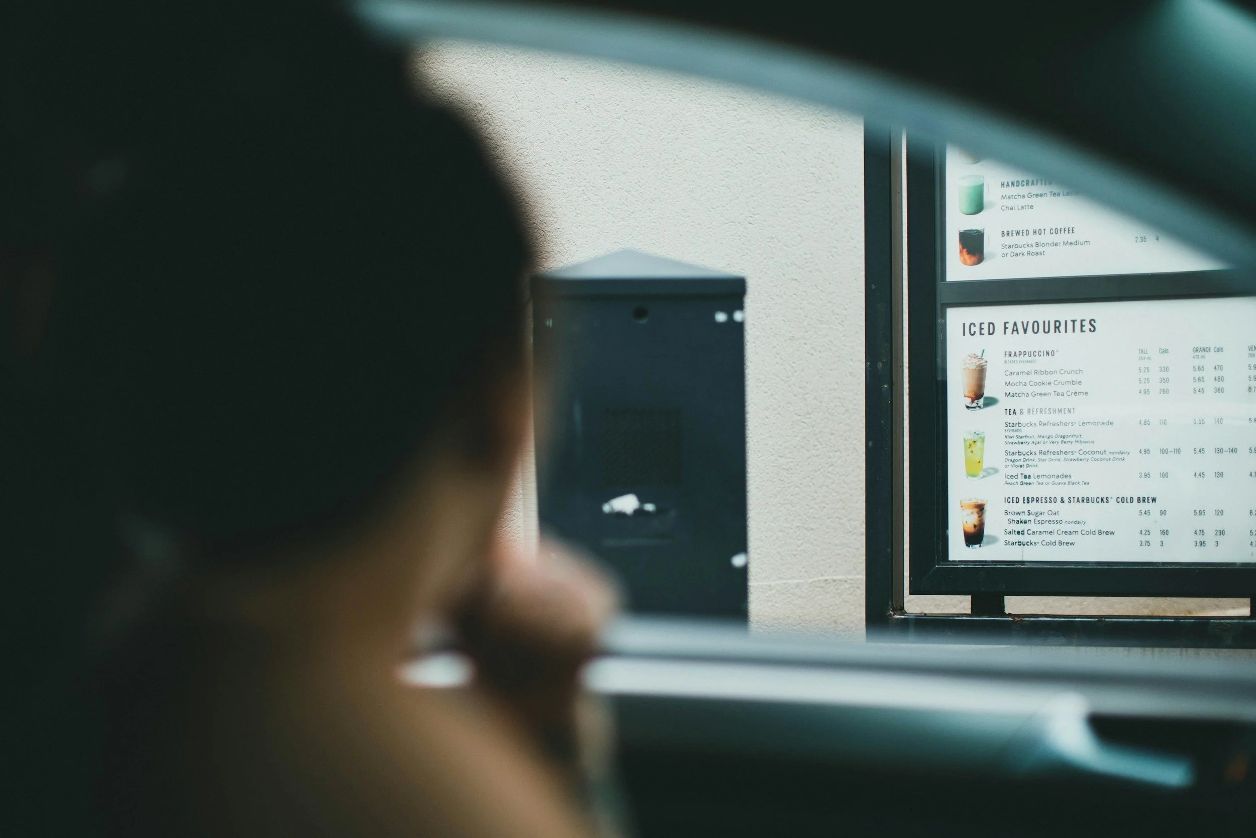A blurry close-up of a person looking at a menu on a fast-food restaurant drive-thru window, with the menu displayed on a digital screen behind the glass.