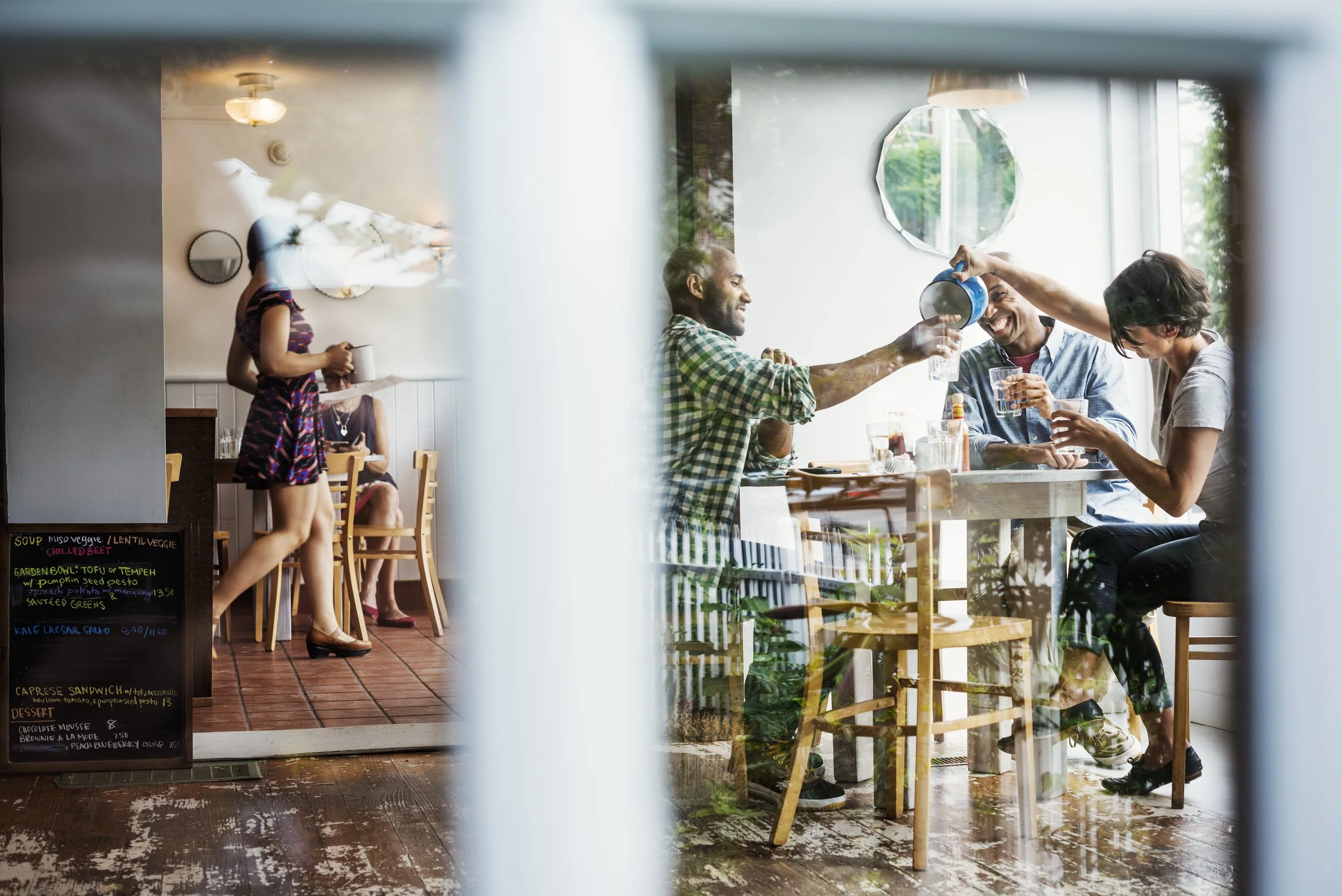 Group of friends enjoying drinks at a restaurant table, with a waitress serving coffee in the background, seen through a window.