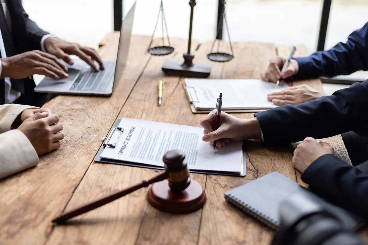 Group of people in suits participating in a contract signing during a business meeting, with legal scales and a gavel on the wooden table.