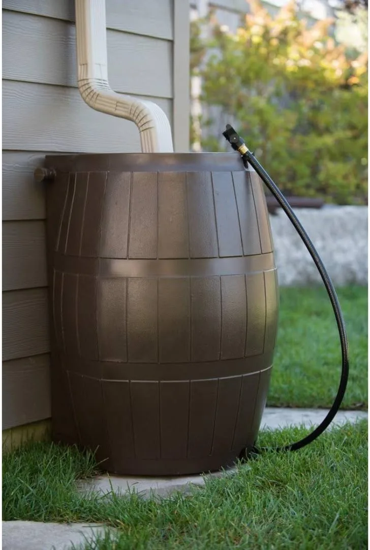 A rain barrel connected to an outdoor downspout with a hose, sitting on a grassy area next to a house with beige siding.