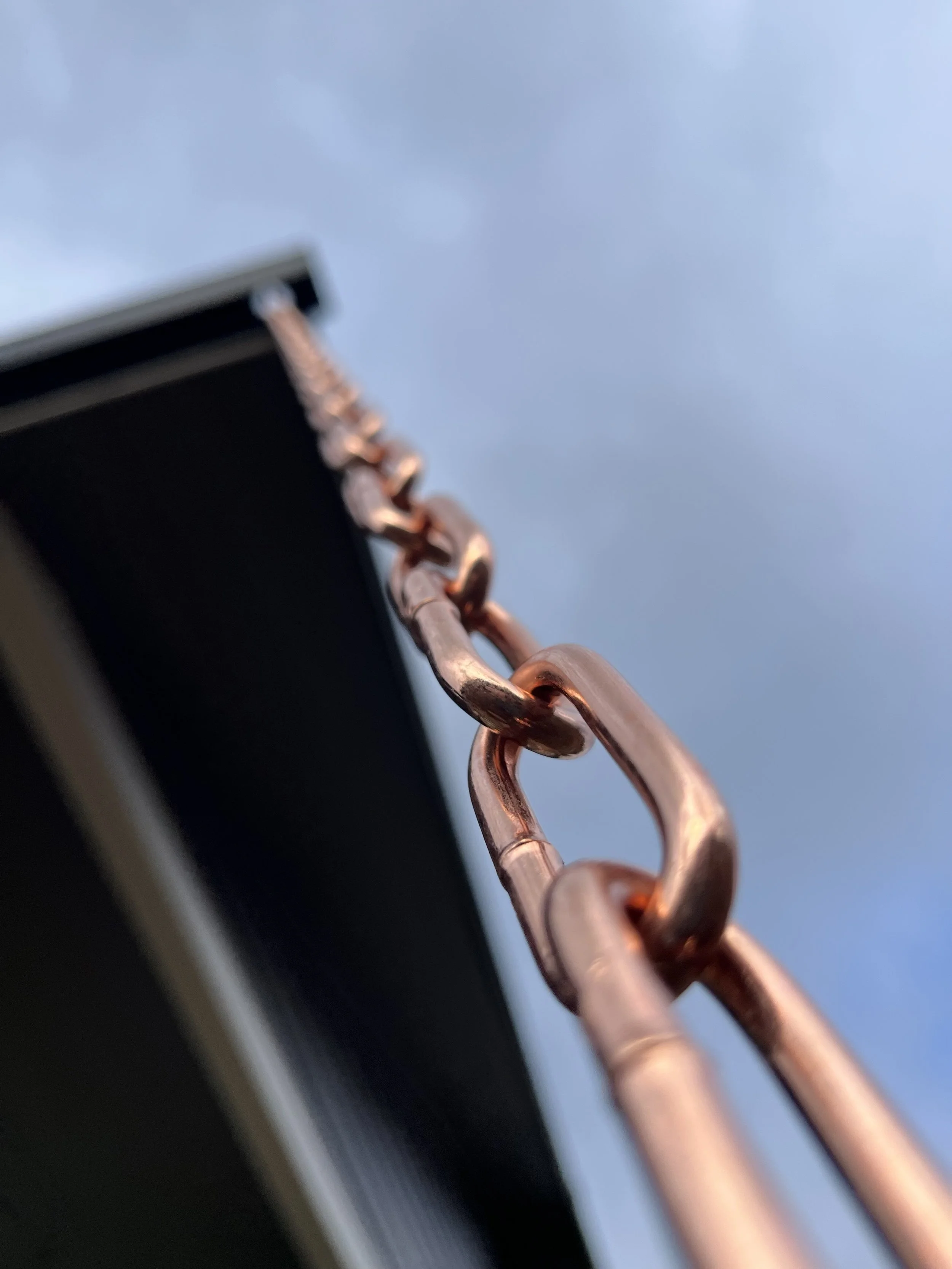 Close-up of a copper chain connected to a black surface, with a blue sky background.