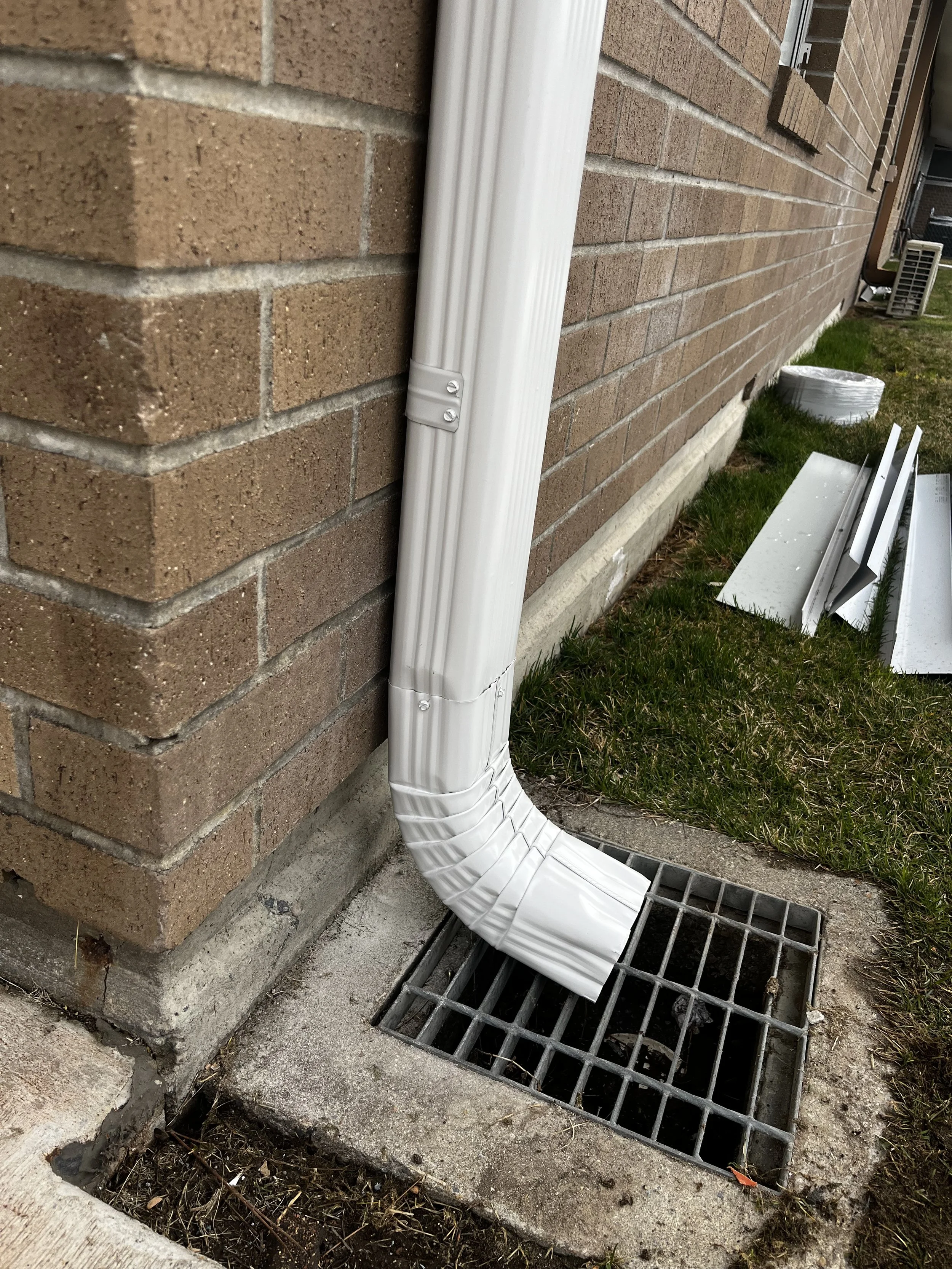 A white plastic downspout extension connected to a rain gutter on the side of a brick house, directing water toward a drain on the concrete ground.