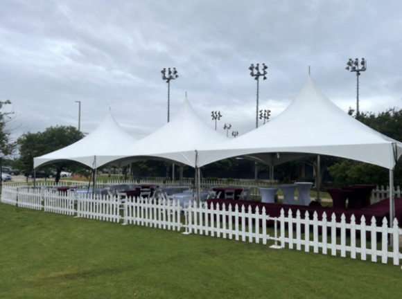Three large white event tents with pointed tops set up on a grassy area with a white picket fence in front, underneath a cloudy sky with tall stadium lights in the background.