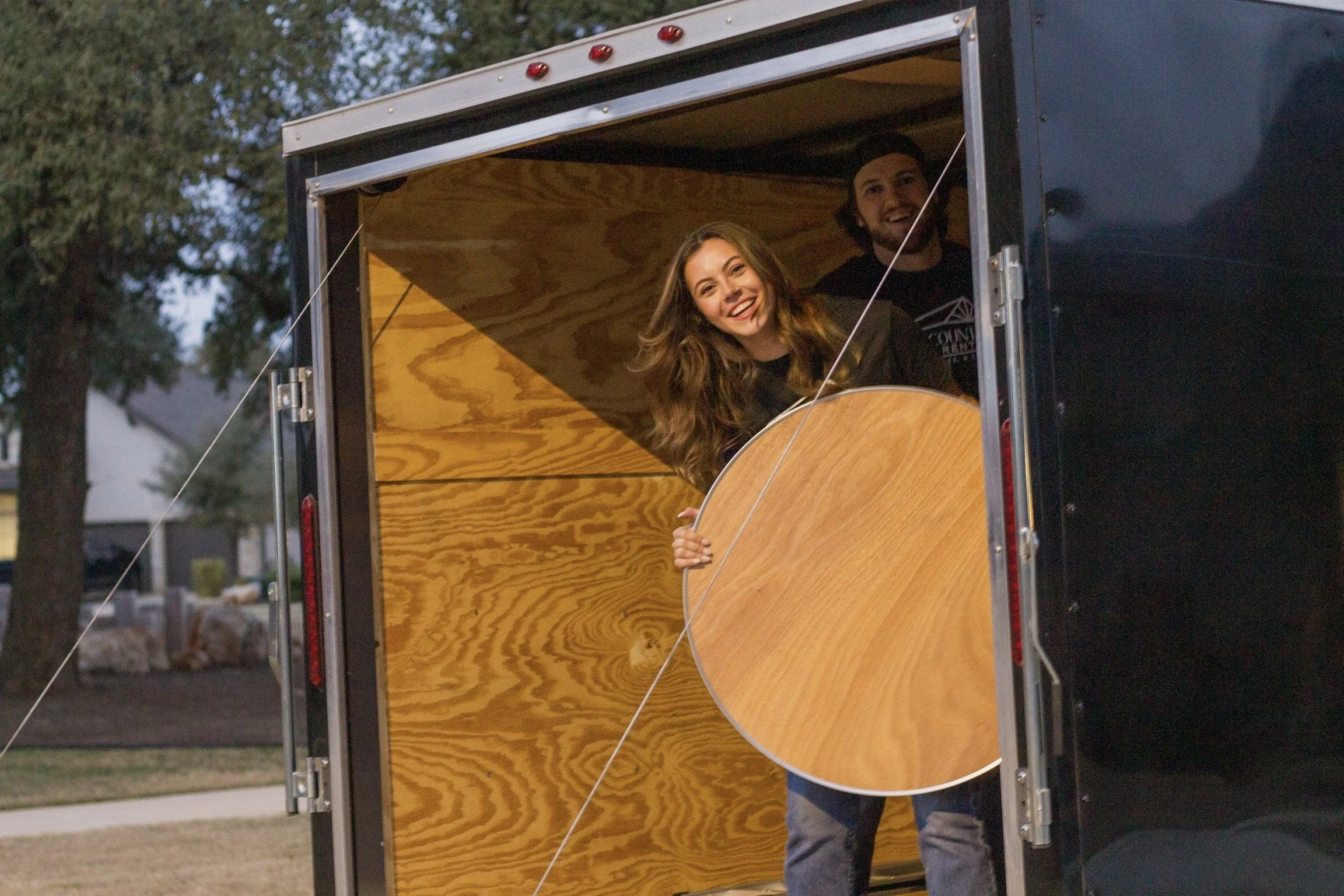 Two people are inside a trailer with a wood interior, one woman holding a guitar and smiling, and a man smiling behind her. The trailer is outdoors with trees in the background.