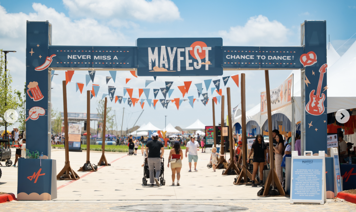 Entrance arch for Mayfest with sign saying 'Never miss a Mayfest, chance to dance!', decorated with patriotic red, white, and blue banners and themed illustrations of musical notes, guitars, and beer mugs, during daytime with people walking past.