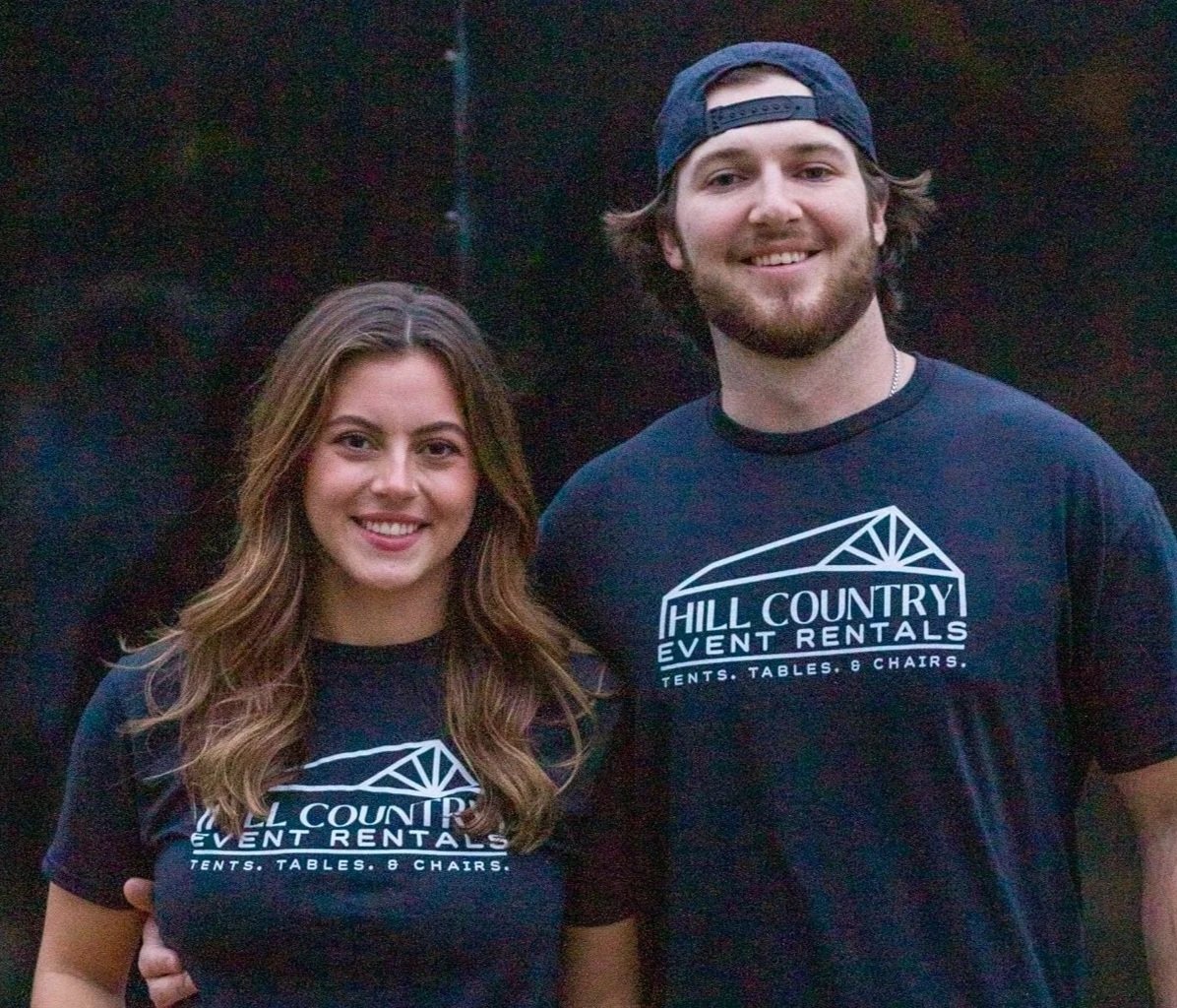 A man and woman smiling and wearing black t-shirts that say Hill Country Event Rentals, outdoors at dusk.