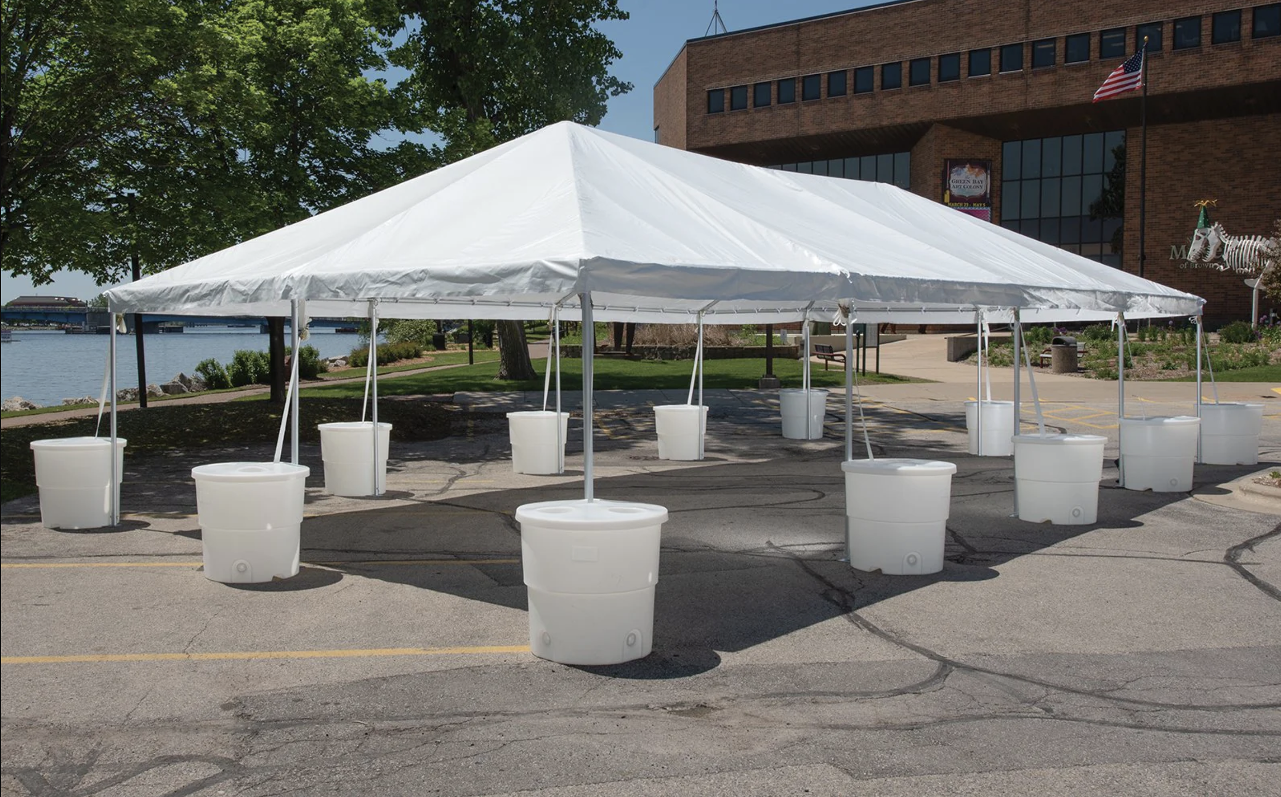 A white event tent set up on a paved parking lot near a river, with trees and a brick building in the background. There are white barrels scattered around the tent.