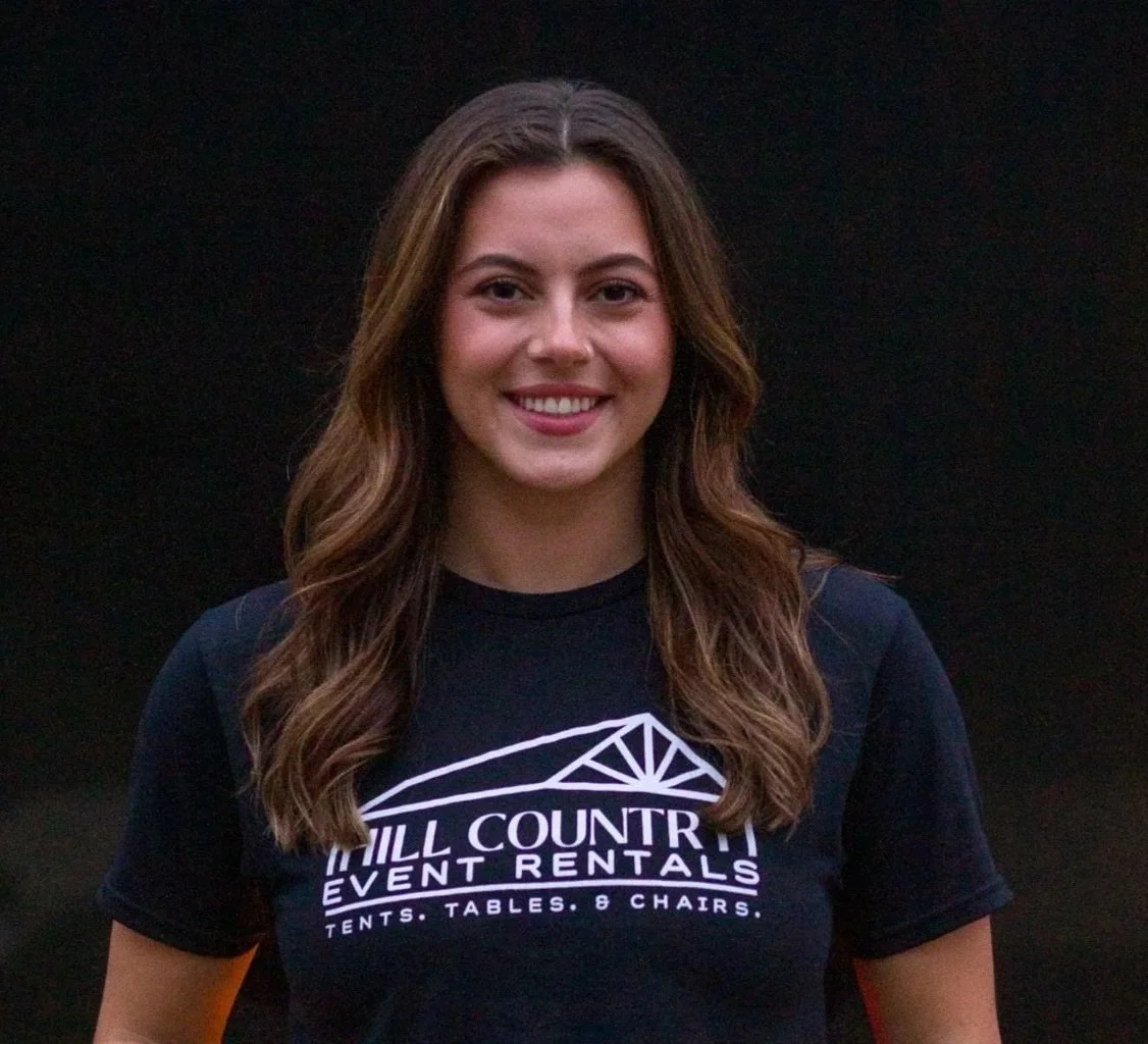 Smiling young woman with wavy brown hair wearing a navy T-shirt with the logo and text 'Hill Country Event Rentals' against a dark background.