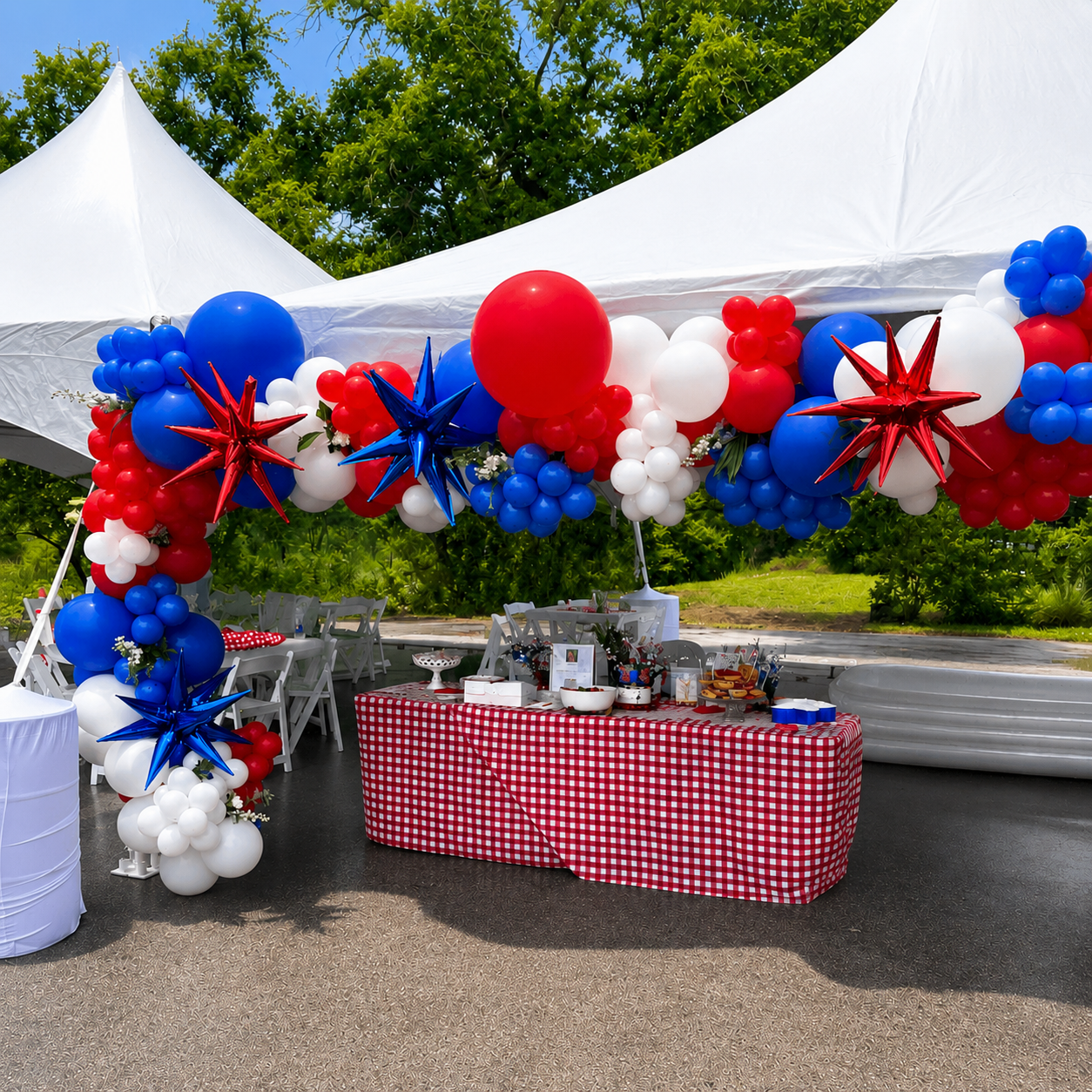Patriotic outdoor celebration decorated with red, white, and blue balloons and star-shaped ornaments under a white tent, with a checkered tablecloth-covered table and greenery in the background.