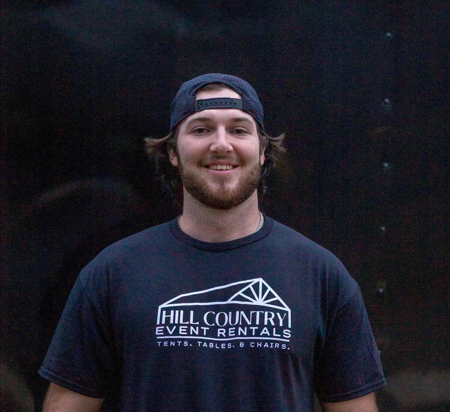 A young man with a beard, wearing a black baseball cap backwards and a dark t-shirt with a logo for Hill Country Event Rentals, standing against a black starry night sky background.