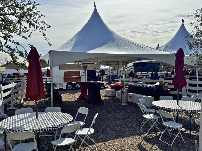 Outdoor event setup with white canopy tent, round tables with checkered tablecloths, folding chairs, and red umbrellas, under partly cloudy sky.
