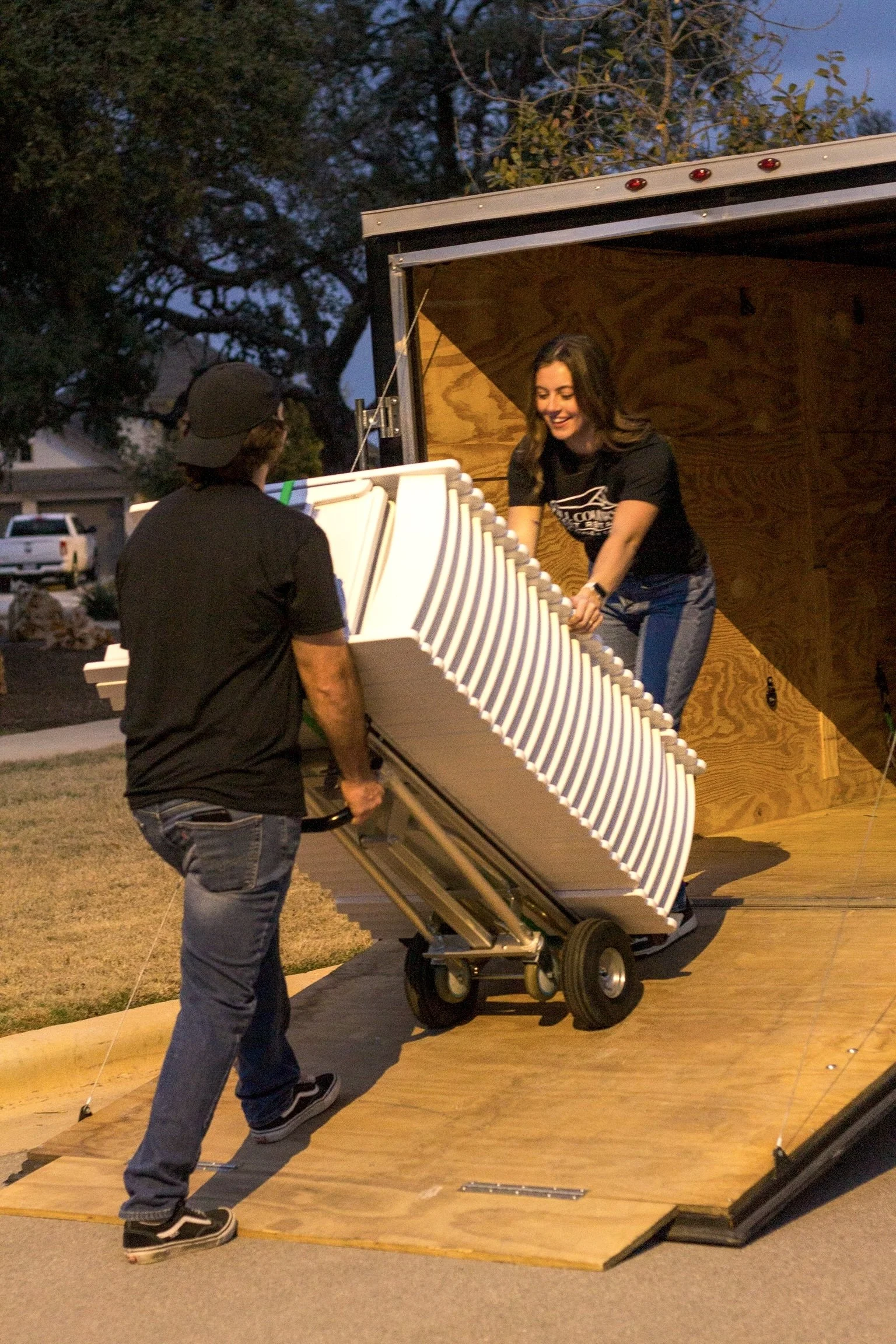 Two people are setting up a large portable piano on a wooden stage outdoors at dusk. One person is pushing a dolly with the piano, while the other is holding the piano and smiling.