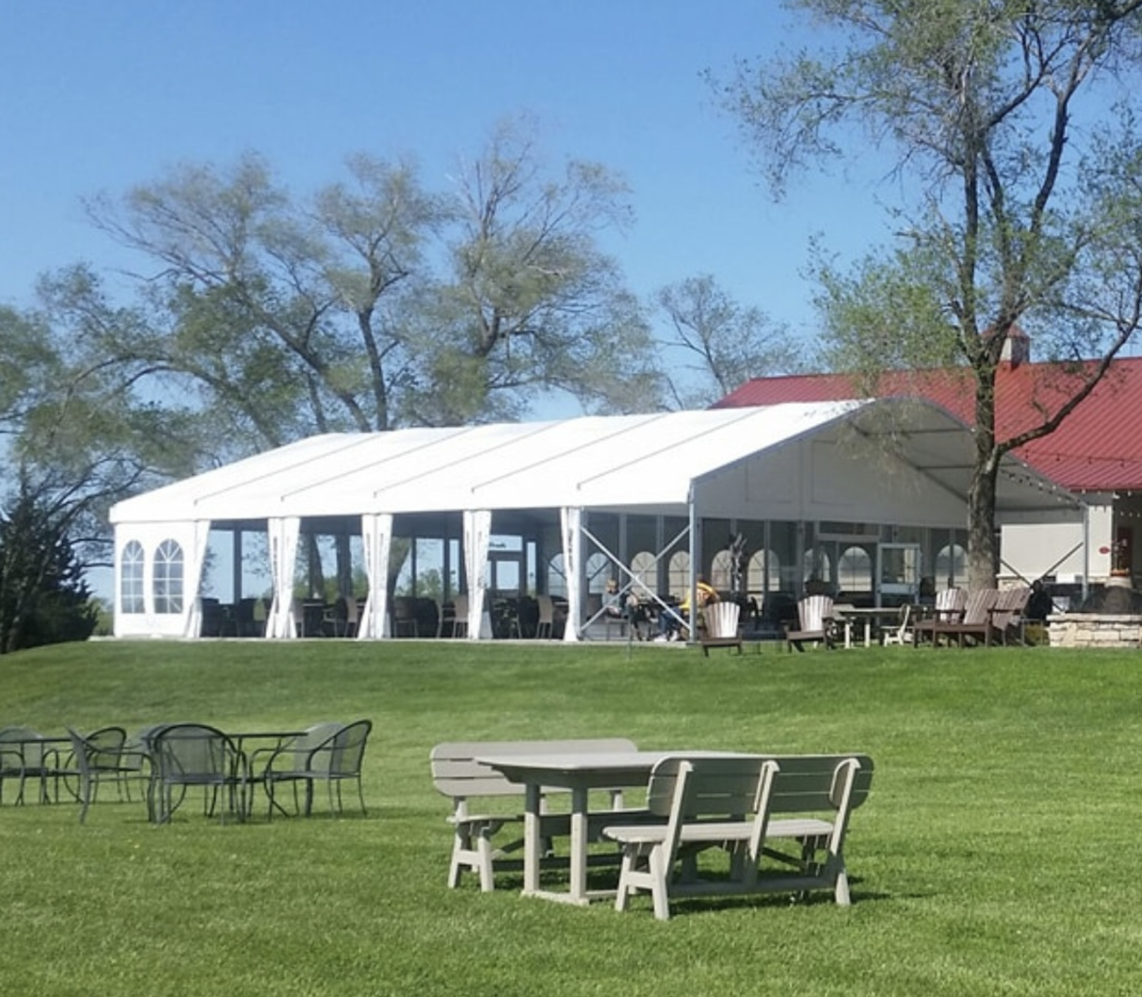 A white event tent set up on a grassy lawn with a red-roofed building and trees in the background, and outdoor chairs and tables in the foreground.