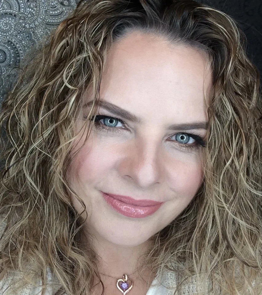 Chris, Close-up of a woman with curly blonde hair, blue eyes, subtle makeup, and a purple heart-shaped pendant necklace.