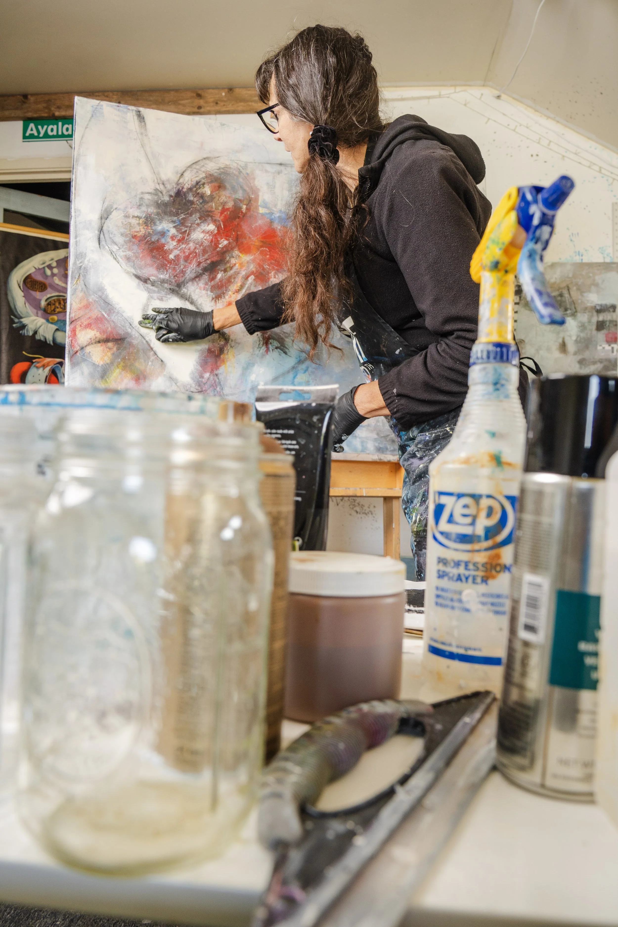 An artist painting an abstract piece on a canvas in a studio, with paint supplies and spray bottles on the table.