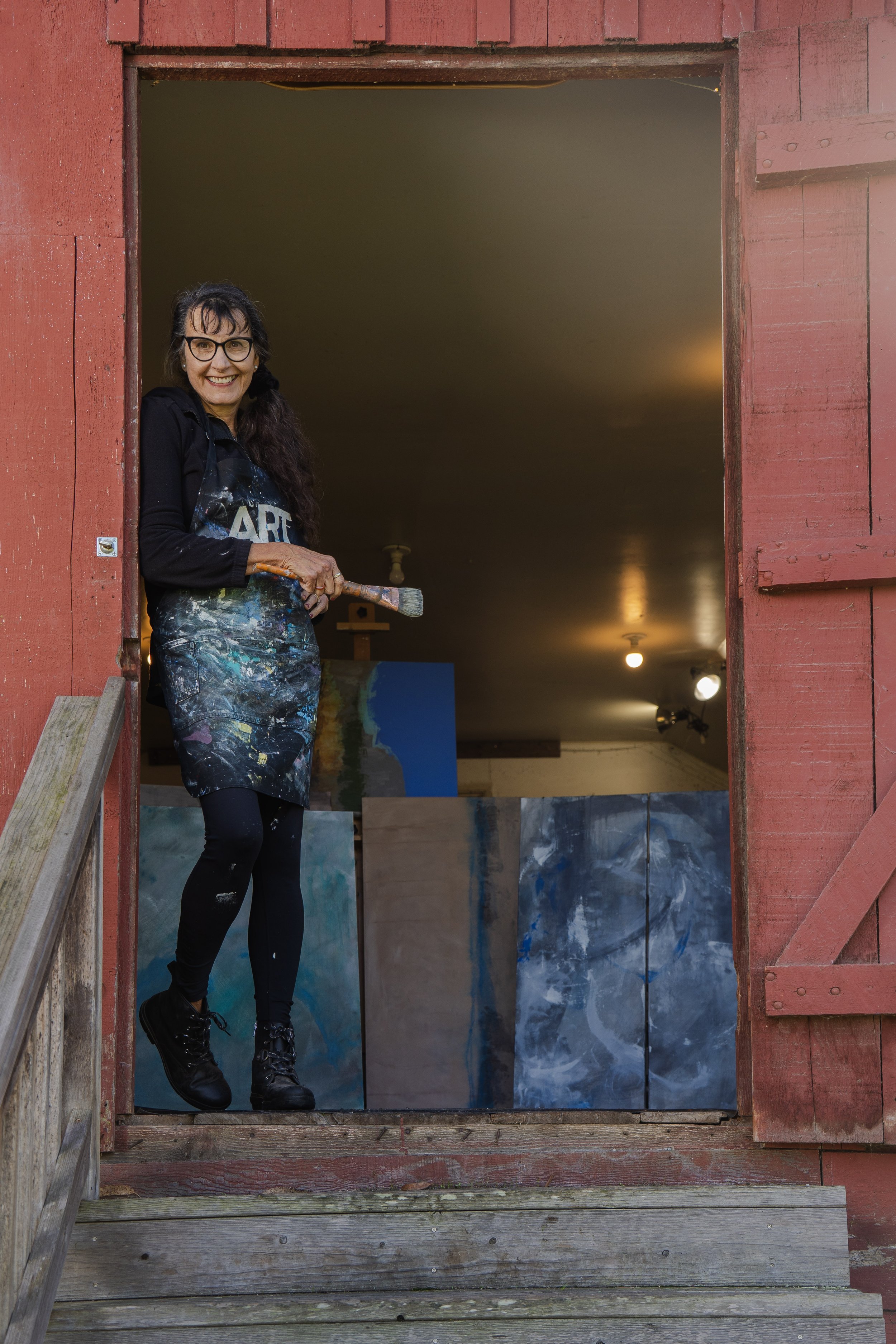 A woman standing in a doorway of a red wooden barn, holding a paintbrush, wearing black glasses, a black jacket, and a paint-stained apron, smiling.