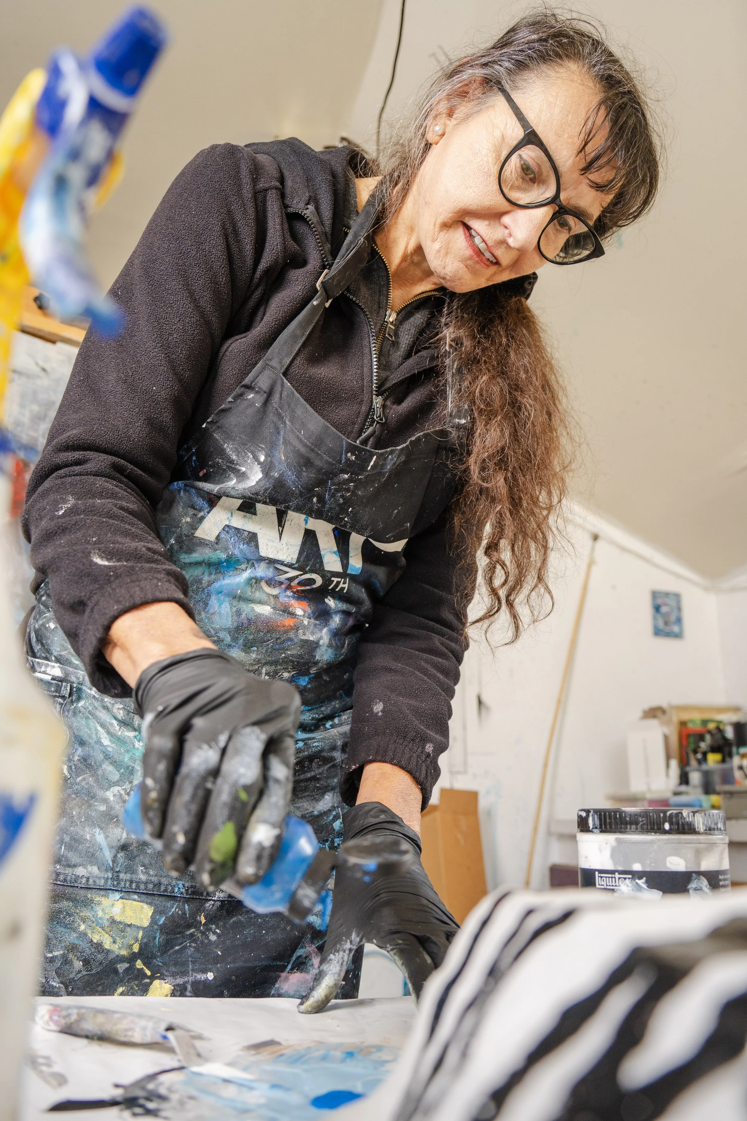 A woman with a paint-splattered apron is working on an art project in a studio. She has long brown hair and appears focused.