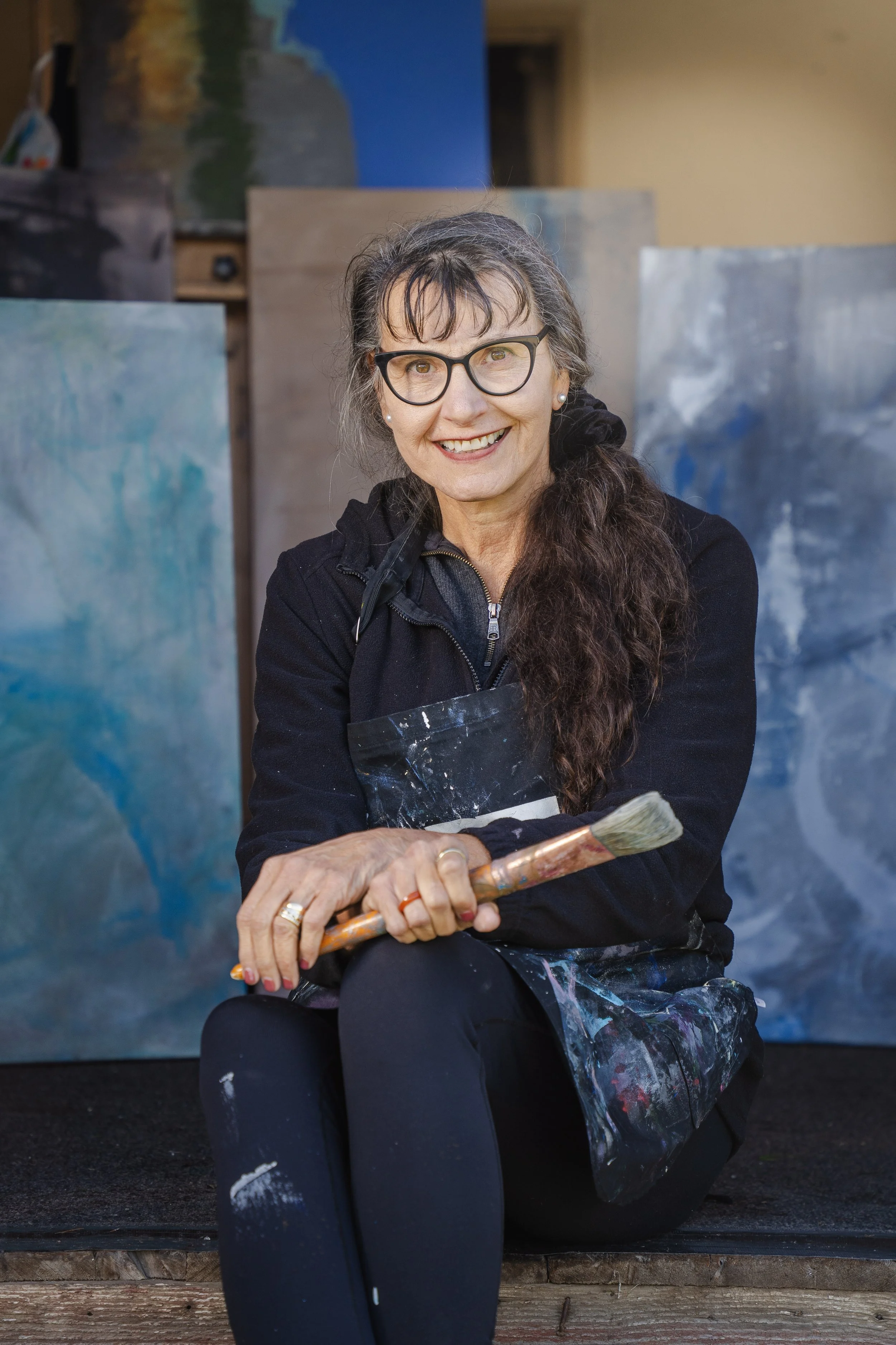 A smiling female artist with long dark hair sitting on the steps of her art studio, holding a paintbrush, with paintings in the background.