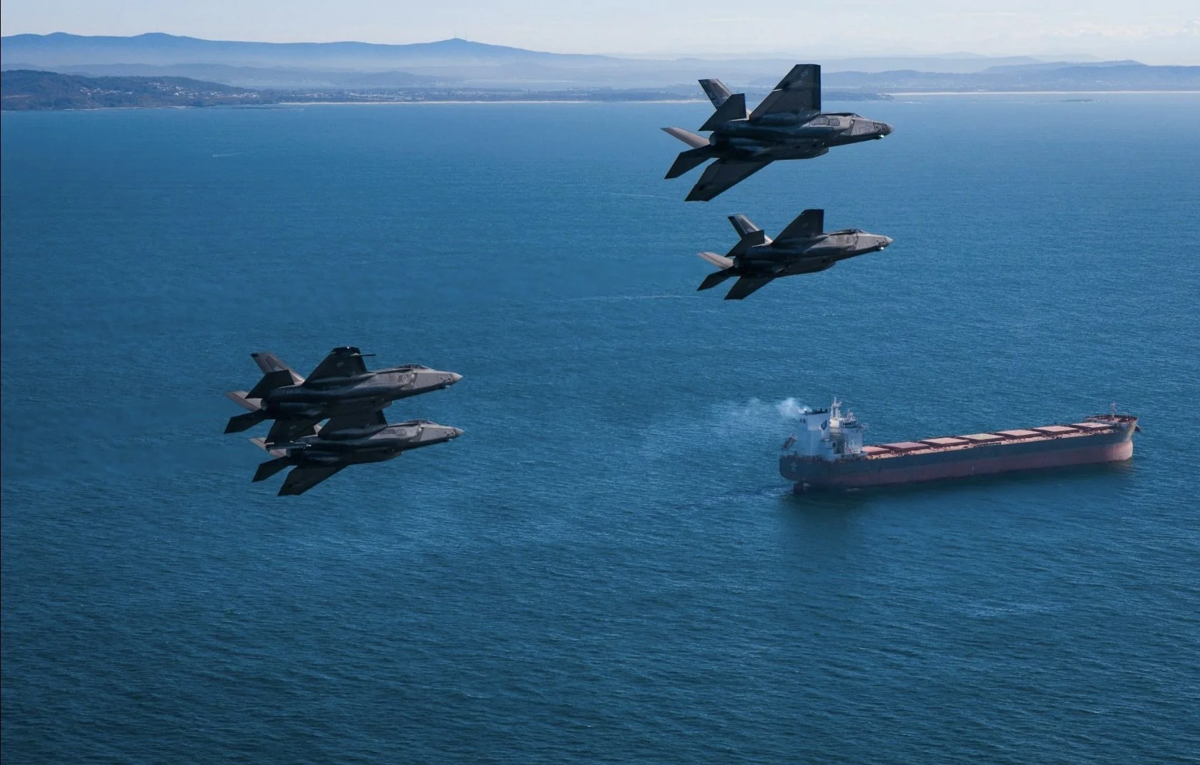 Four fighter jets flying in formation over the ocean with a cargo ship below.