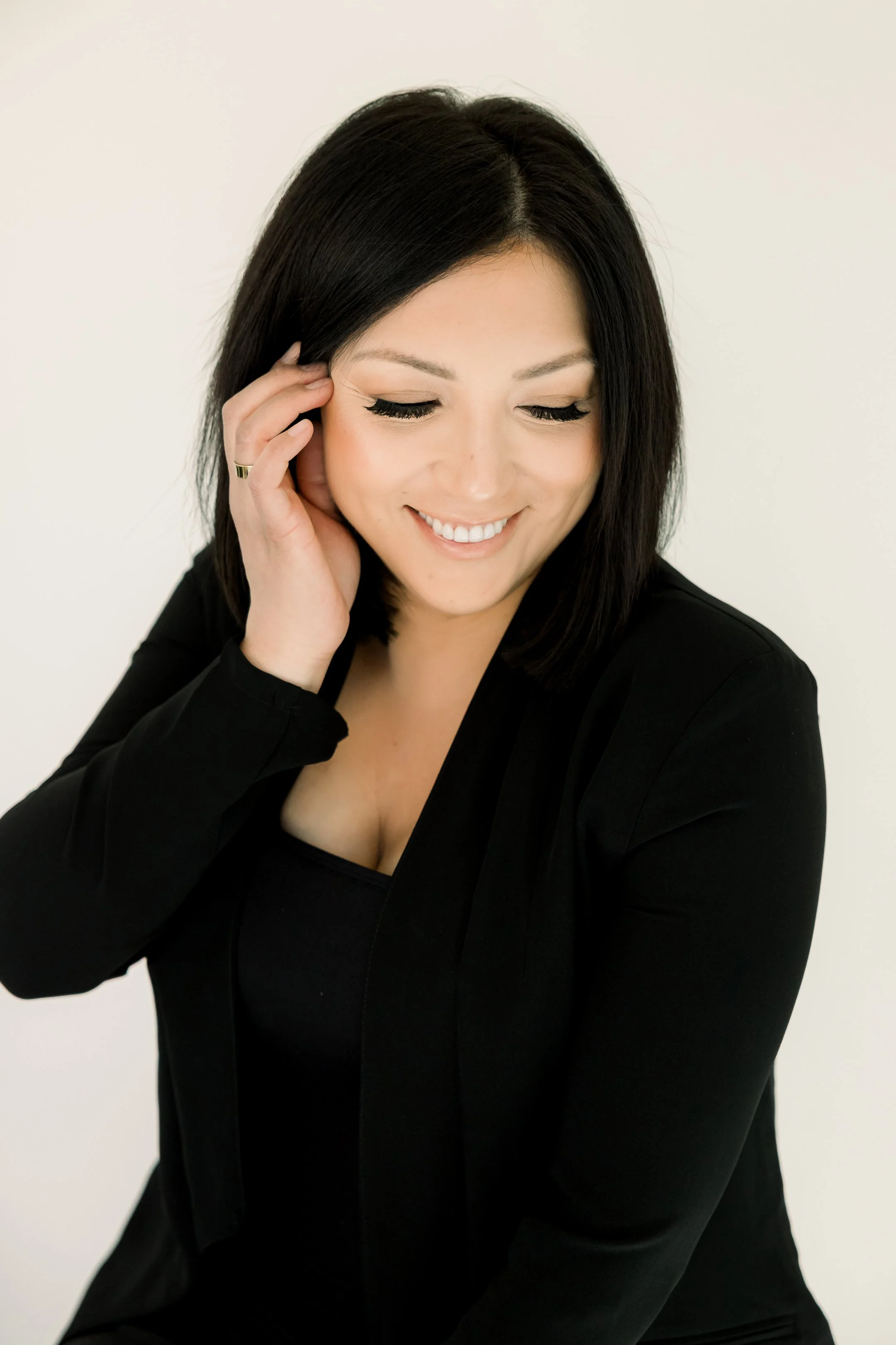 A woman with black hair smiling, wearing a black blazer and top, touching her face with her left hand.