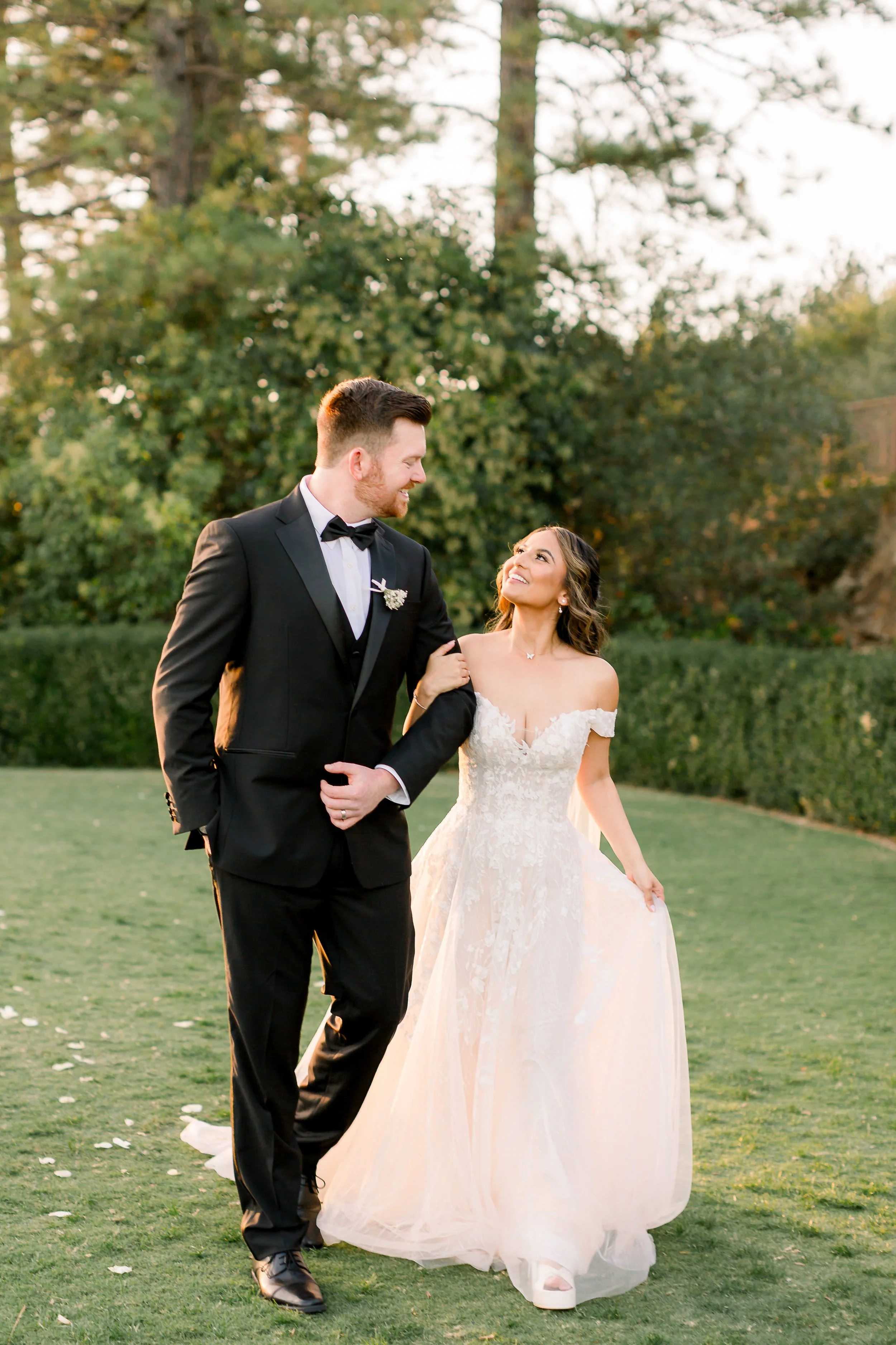A bride and groom walking arm in arm on a grassy lawn during sunset, with trees and bushes in the background, smiling and looking at each other.